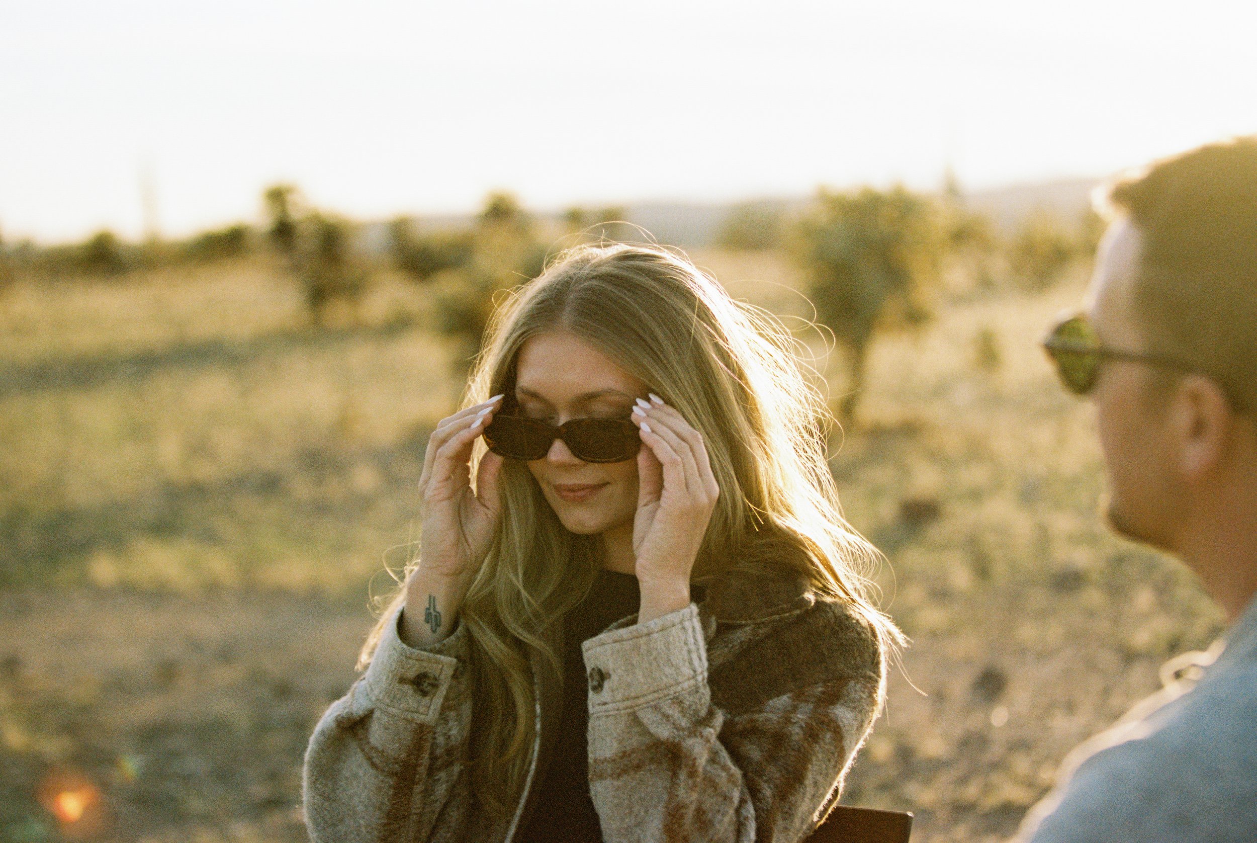 A woman with long blonde hair and sunglasses adjusting her sunglasses outdoors during sunset, with a man wearing sunglasses in the background, in a desert landscape.