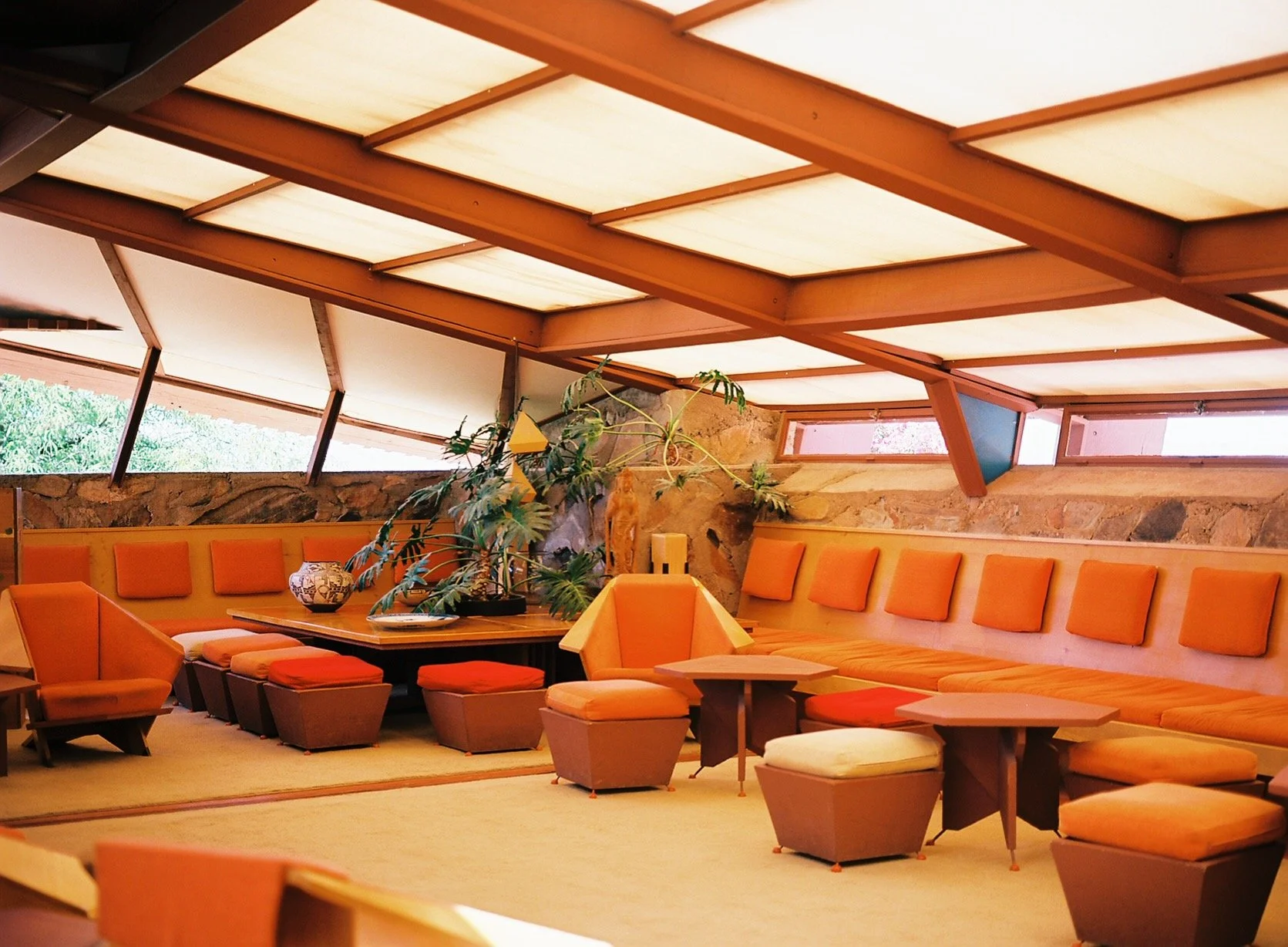 A mid-century modern lounge with orange cushioned seating, multiple small wooden tables, a large green plant, and a stone wall with skylights in the ceiling.