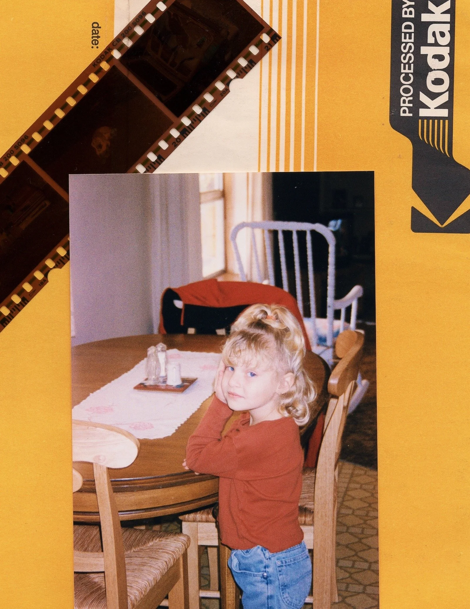 A young girl with blond curly hair sitting at a wooden dining table, looking at the camera with her head resting on her hand. The table has a white placemat and salt and pepper shakers. Behind her, there's a chair, a window, and various household ite