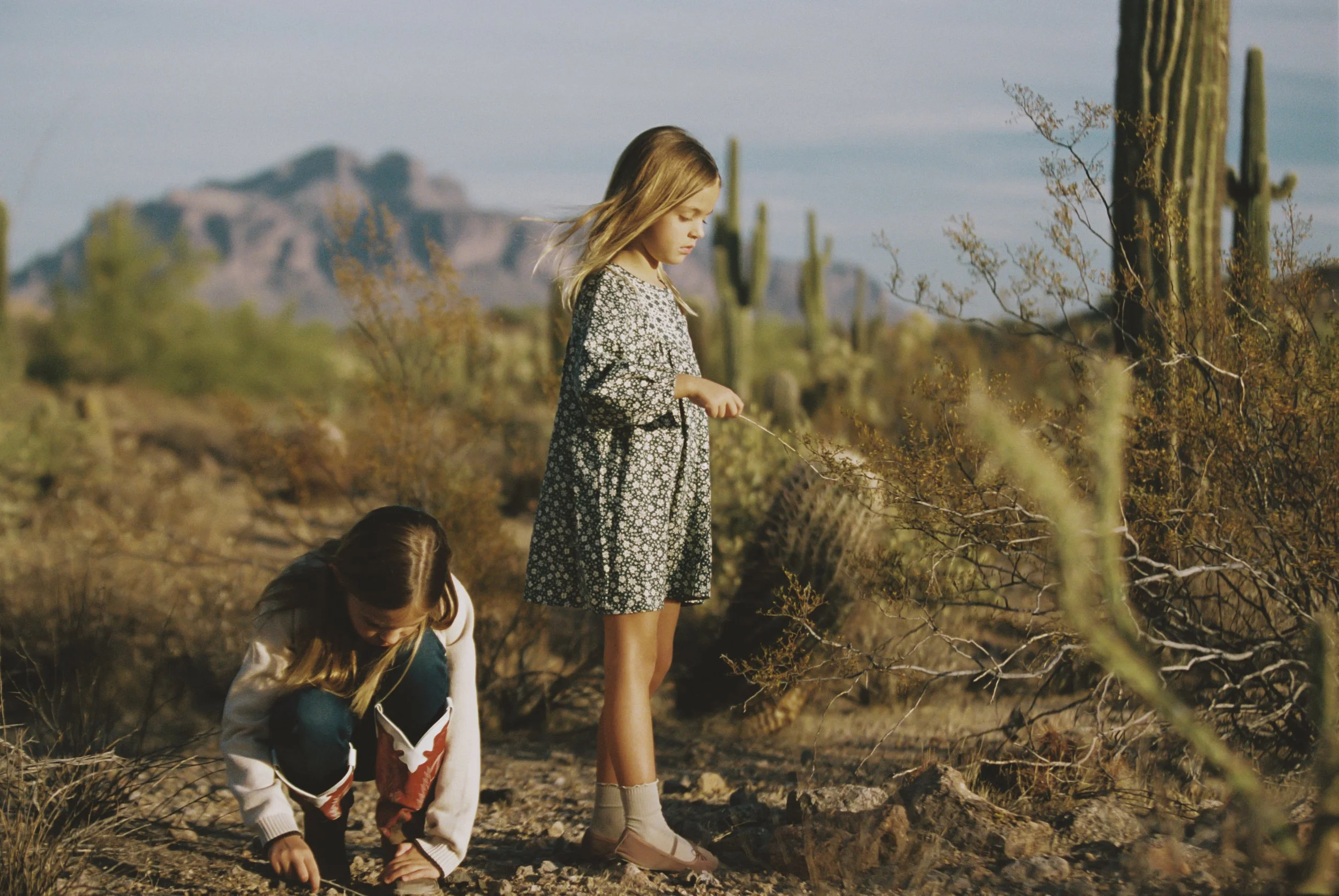 Two young girls in a desert landscape with cacti and mountains in the background. One girl is standing and examining a plant, while the other girl is crouching and picking something from the ground.