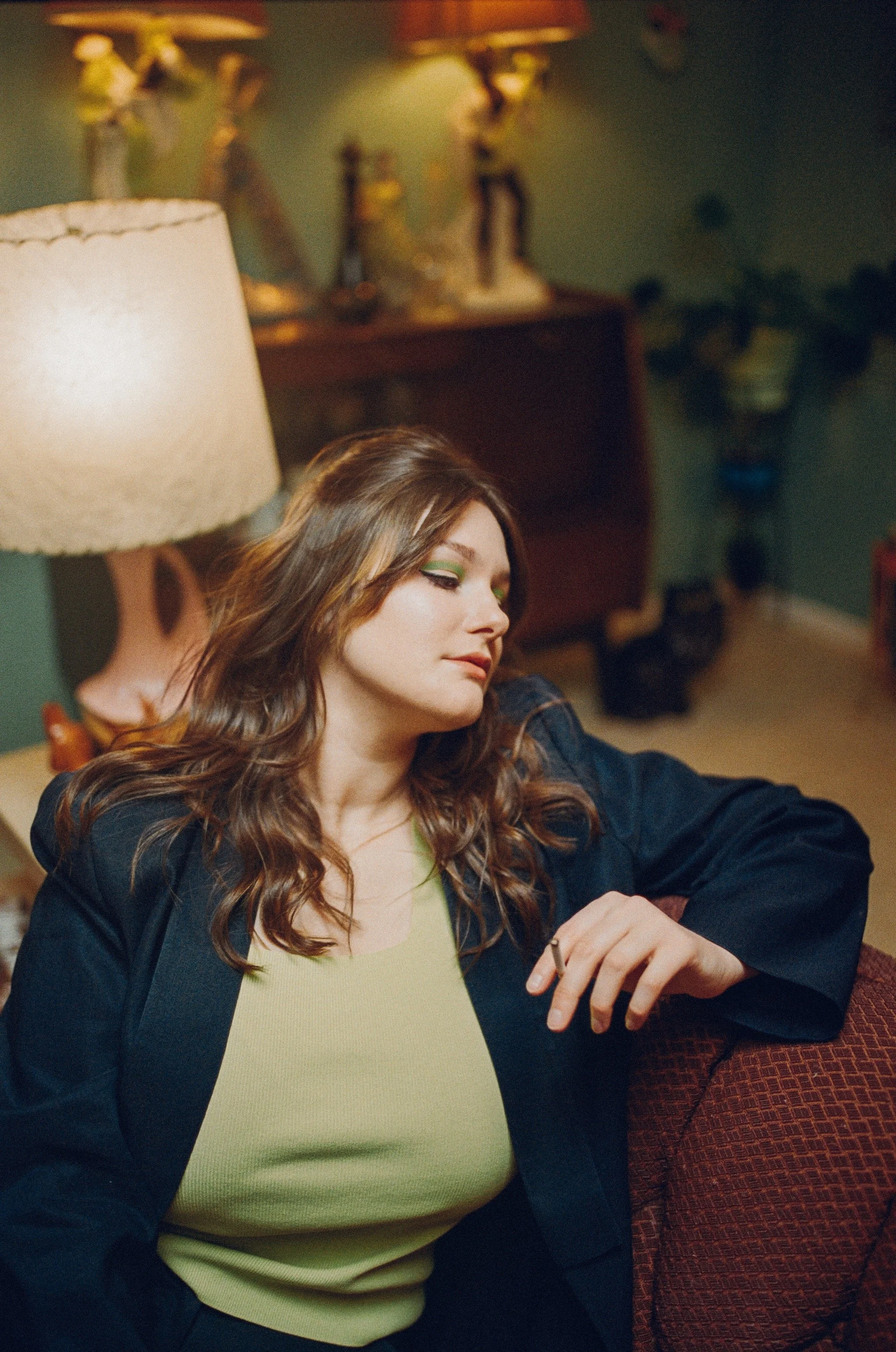 A woman with wavy brown hair and green eyeshadow sitting on a red patterned chair in a room with vintage decor, including a lamp, wooden furniture, and decorative figurines in the background.