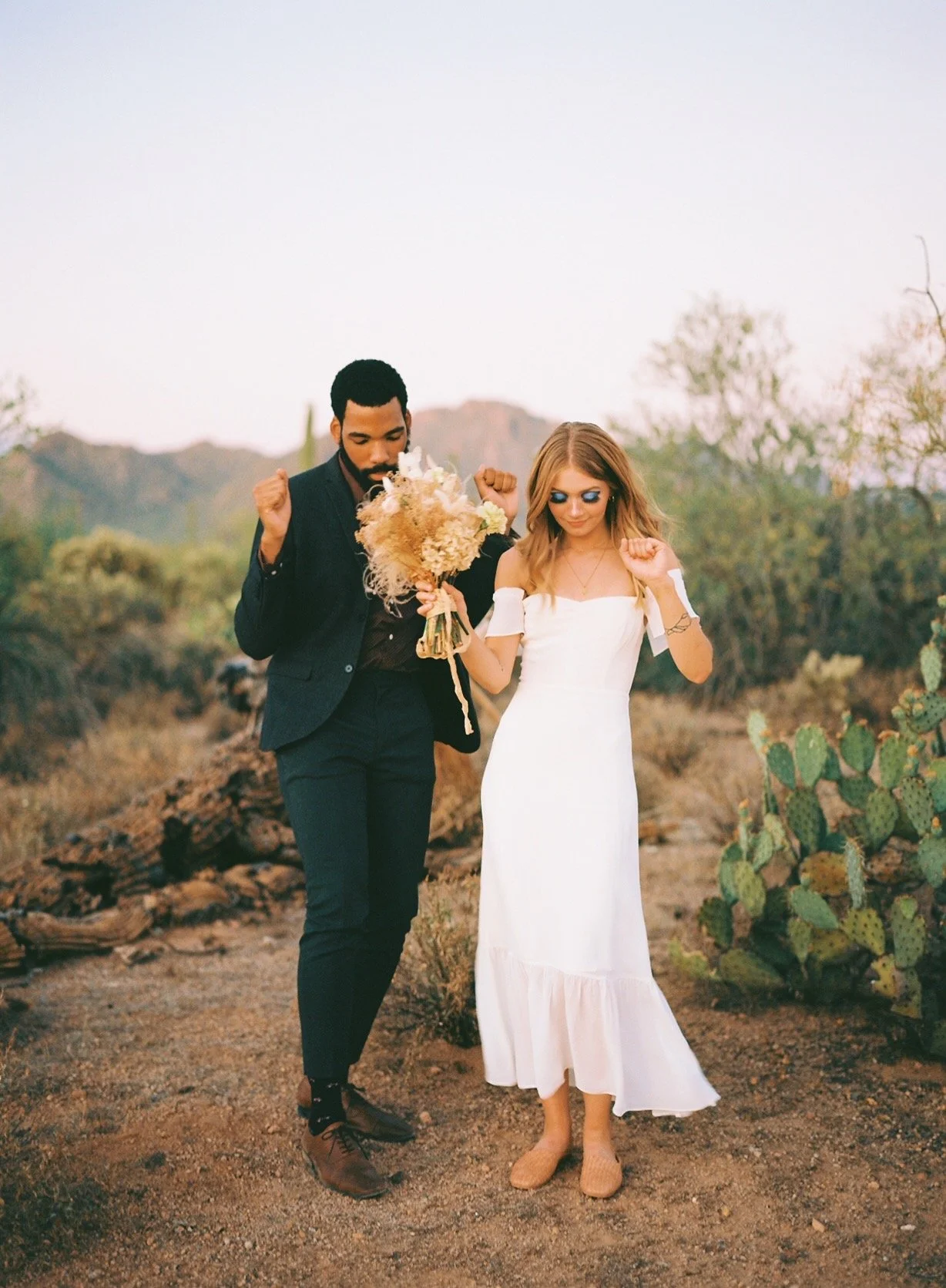 A couple dancing together outdoors in a desert landscape, with cacti and rocky hills in the background. The woman is wearing a white dress and holding a bouquet, and the man is dressed in a dark suit. They look happy and relaxed.
