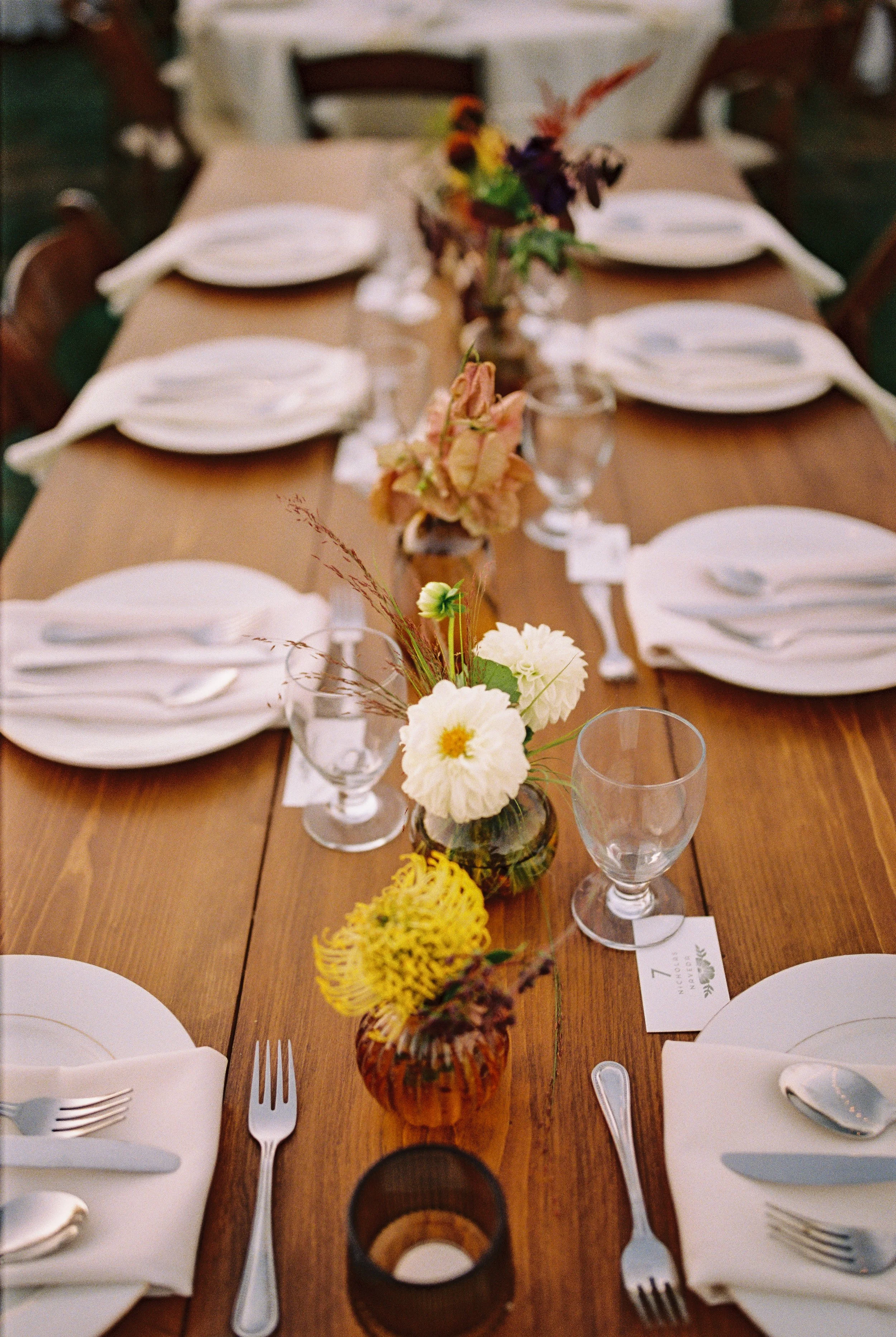 A rustic wooden dining table set for a meal with white plates, silverware, and clear water glasses. The table features a floral centerpiece in various small vases with colorful flowers and greenery.
