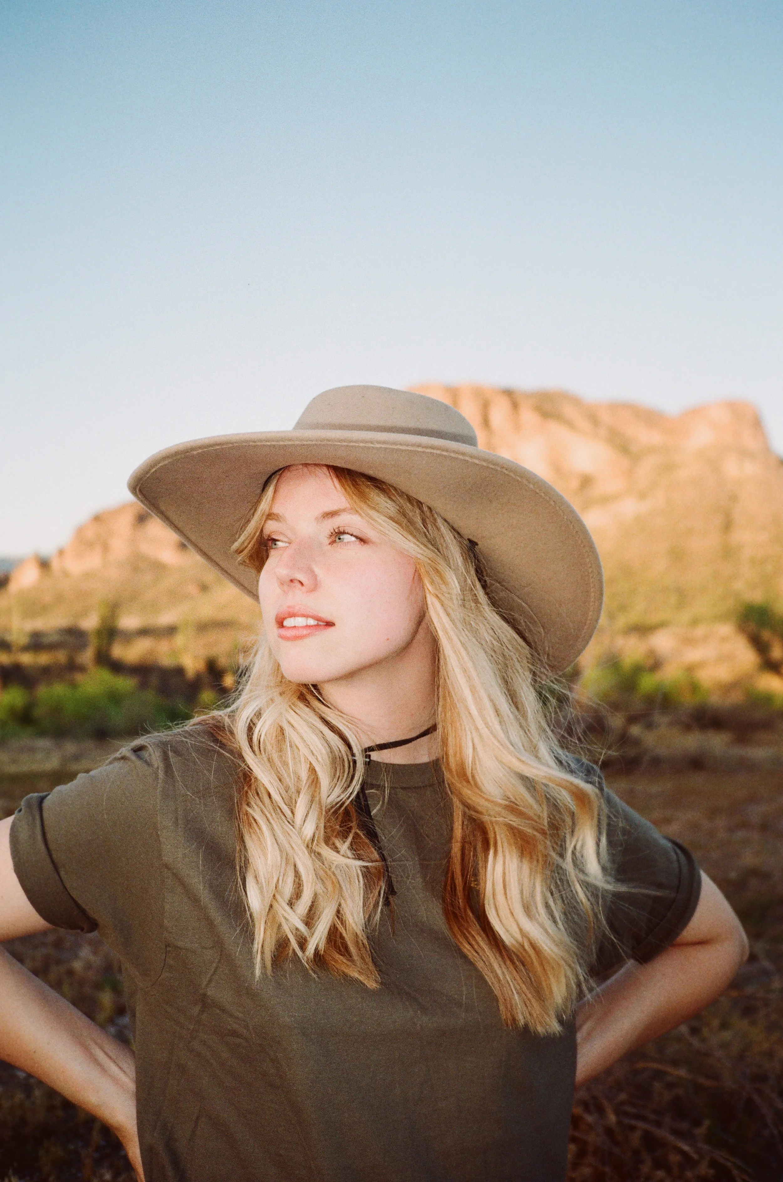 A young woman with blonde hair wearing a wide-brimmed hat and a dark green t-shirt standing outdoors in a desert landscape with rock formations in the background during sunset.