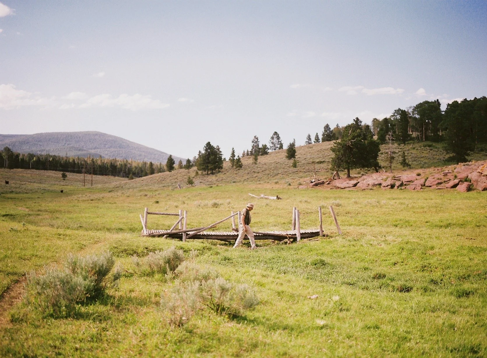 A man walking across a small wooden bridge in a grassy open field with hills, trees, and mountains in the background.