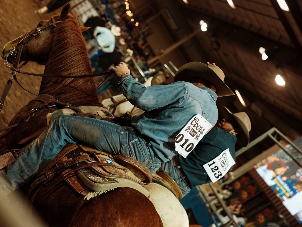 Two cowboys riding horses in an indoor arena during an auction, wearing jeans, cowboy hats, and auction tags.