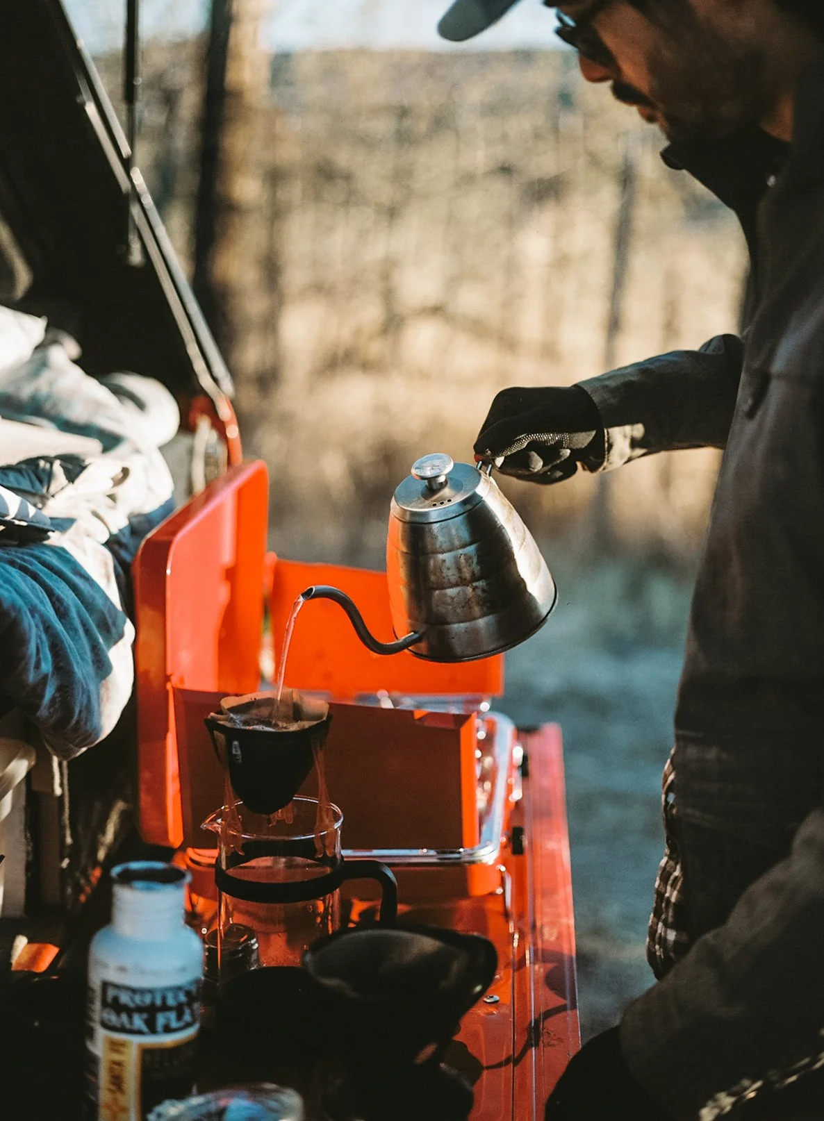 A person pouring hot water from a kettle into a coffee drip cone on a camping stove, with a bright orange box and some supplies nearby, outdoors in a natural setting.