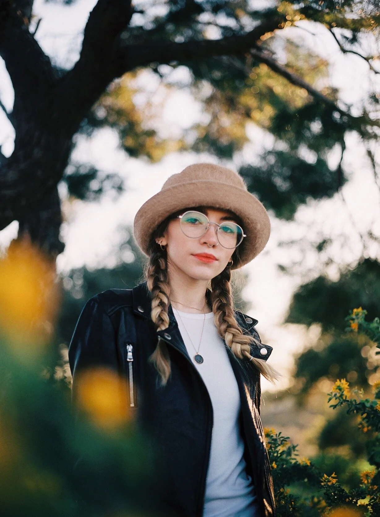A young woman outdoors, wearing a beige felt hat, glasses, a black leather jacket, and a white shirt, standing among yellow flowers with a large tree in the background during golden hour.