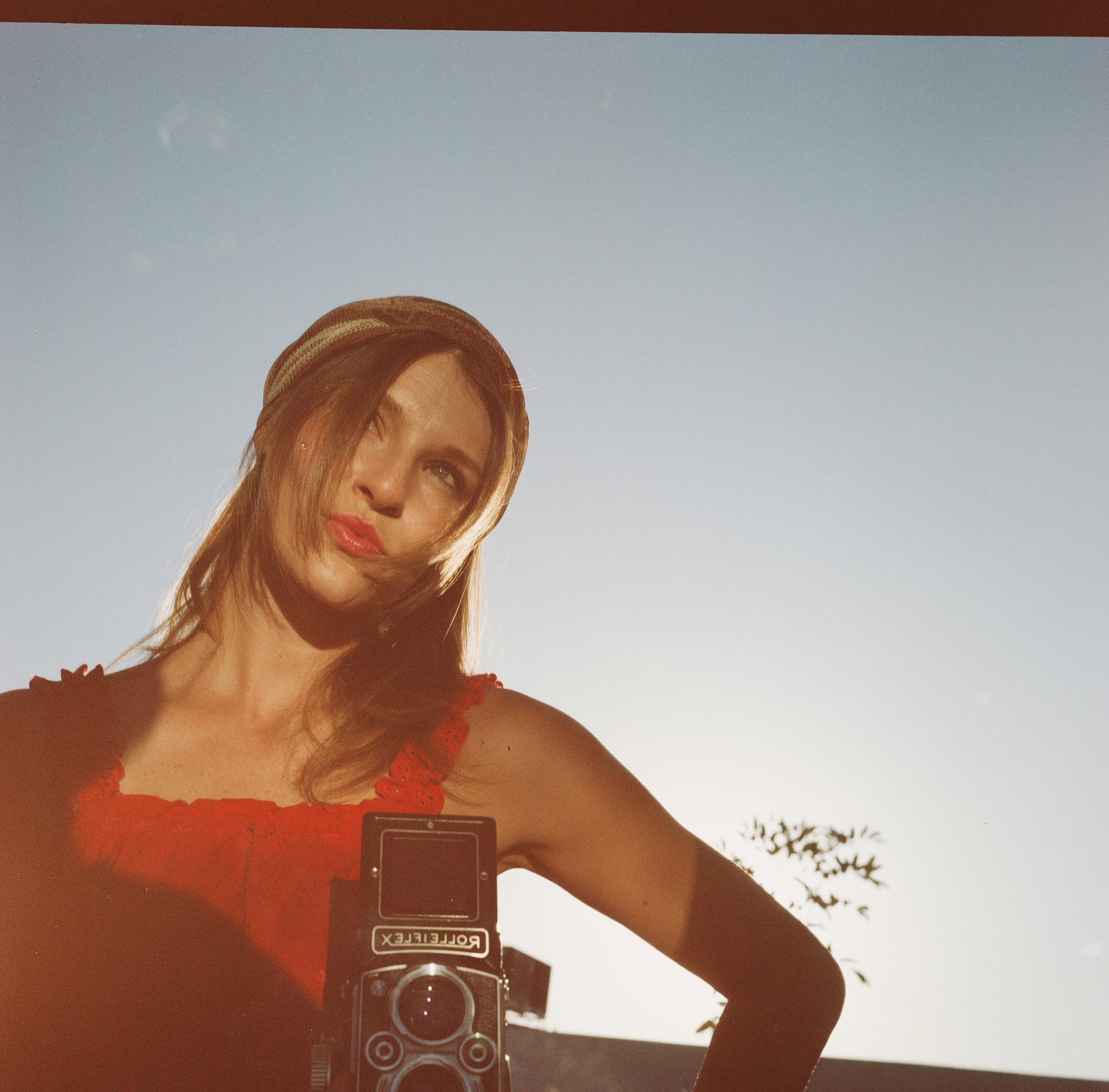 A woman with long hair wearing a red tank top and a tan hat, taking a selfie with a vintage Rolleiflex camera in a mirror outdoors during sunset or sunrise.