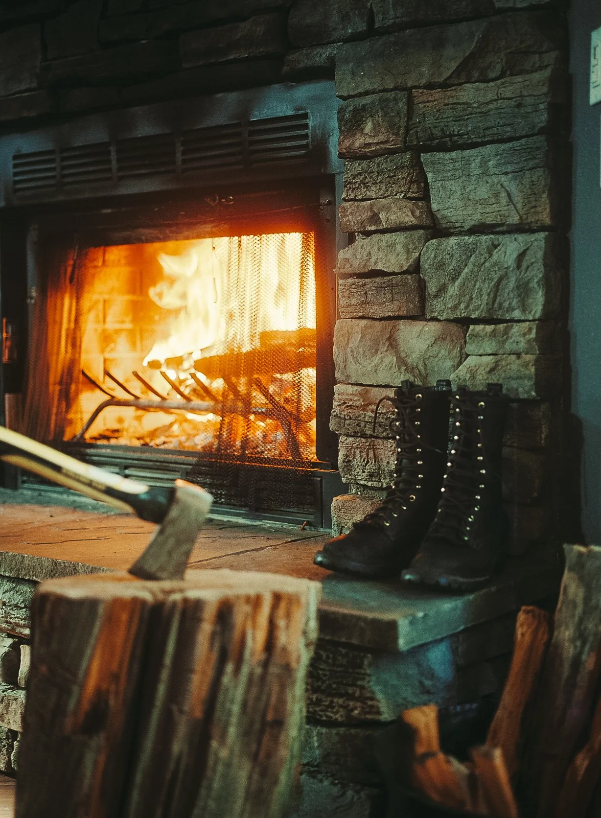 A roaring fireplace with flames and burning logs, a pair of black lace-up boots resting on a wooden hearth, and an axe embedded in a chopped wooden log nearby.