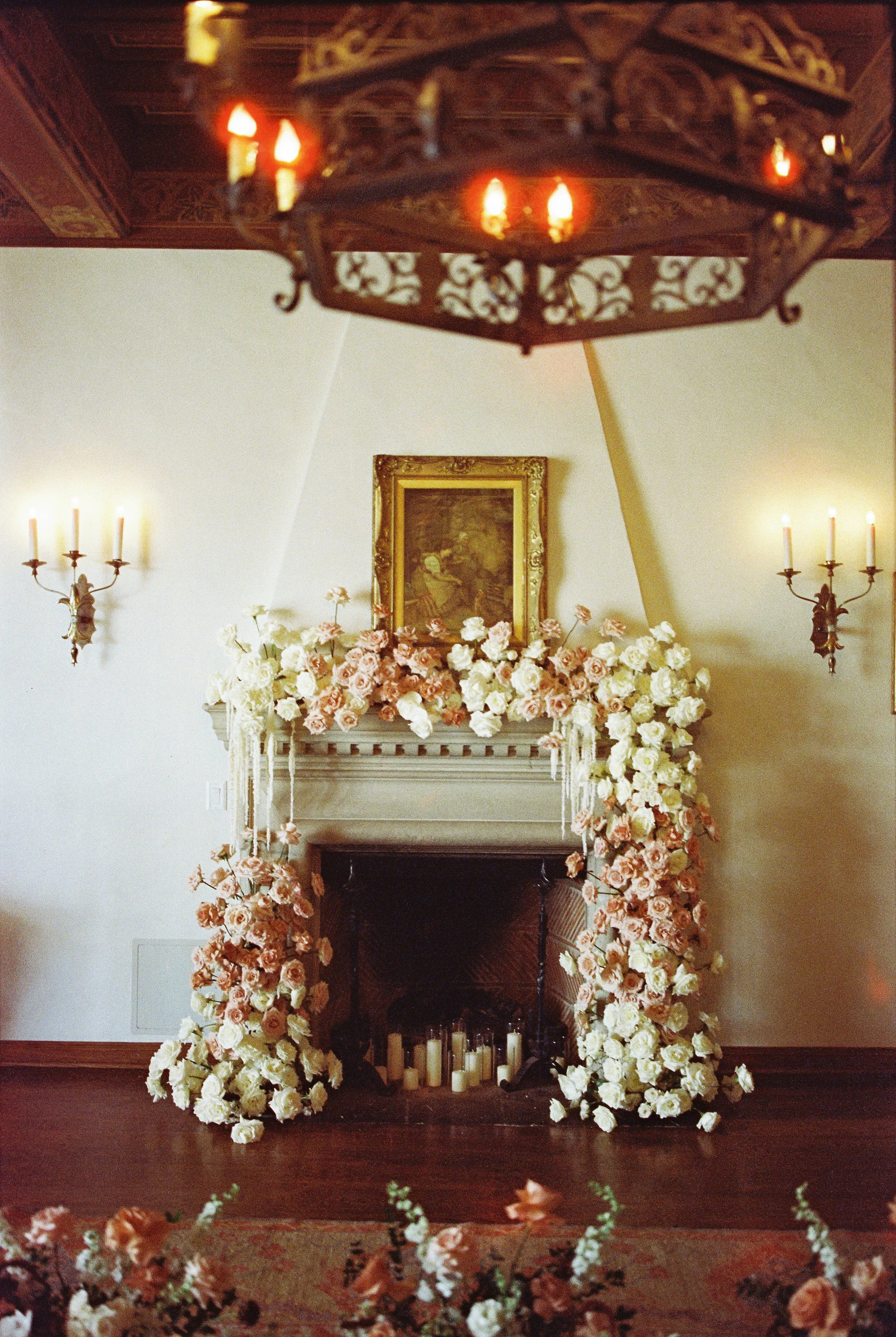 A fireplace decorated with an arrangement of white and pink roses and greenery, with candles inside the fireplace. An ornate gold-framed painting hangs above and two wall-mounted candle sconces flank the fireplace.