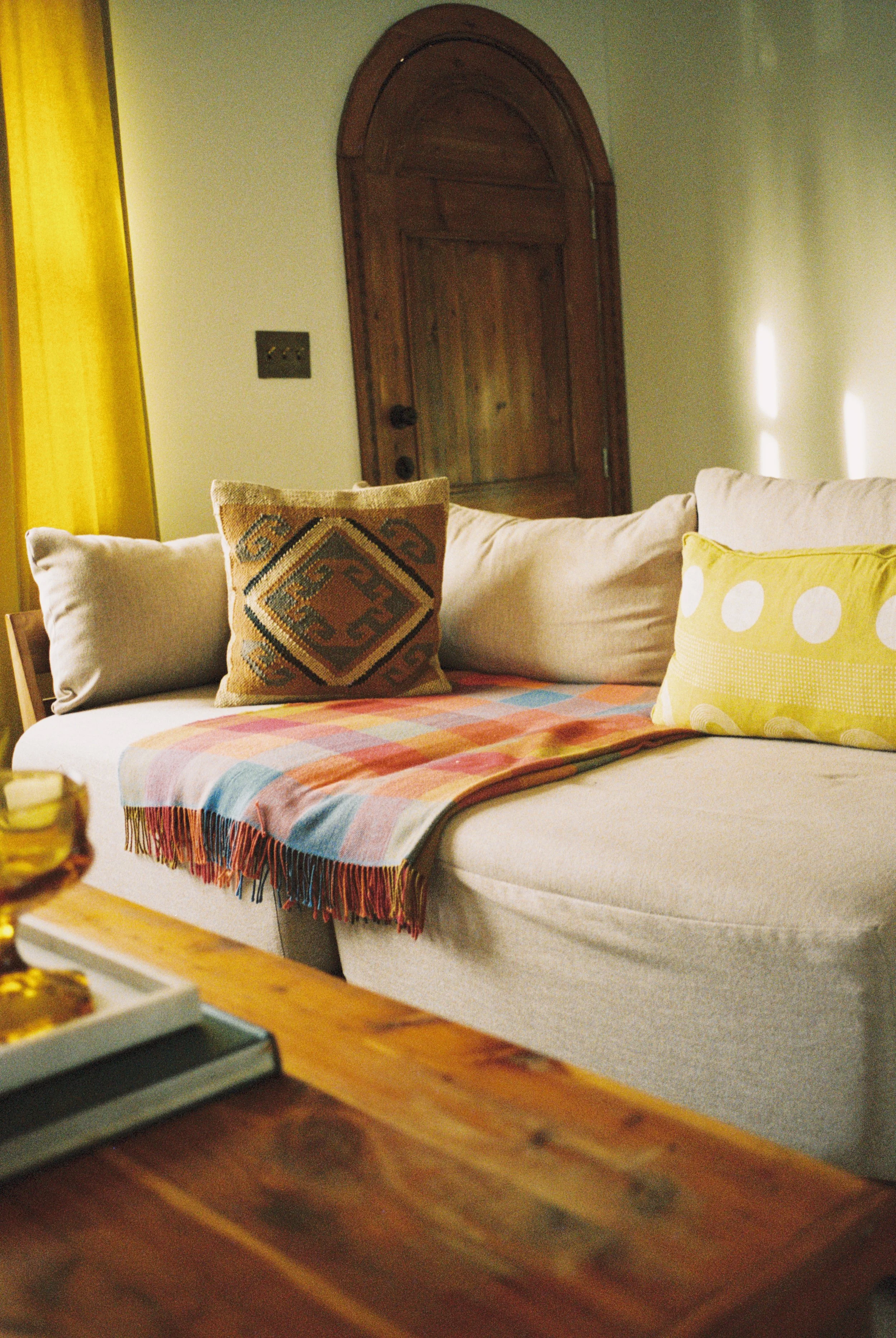 A cozy living room with a beige sofa and colorful throw pillows, a fringed checkered blanket, and a wooden table with a tray of snacks. There's a wooden arched door and yellow curtains in the background, with sunlight casting shadows on the wall.