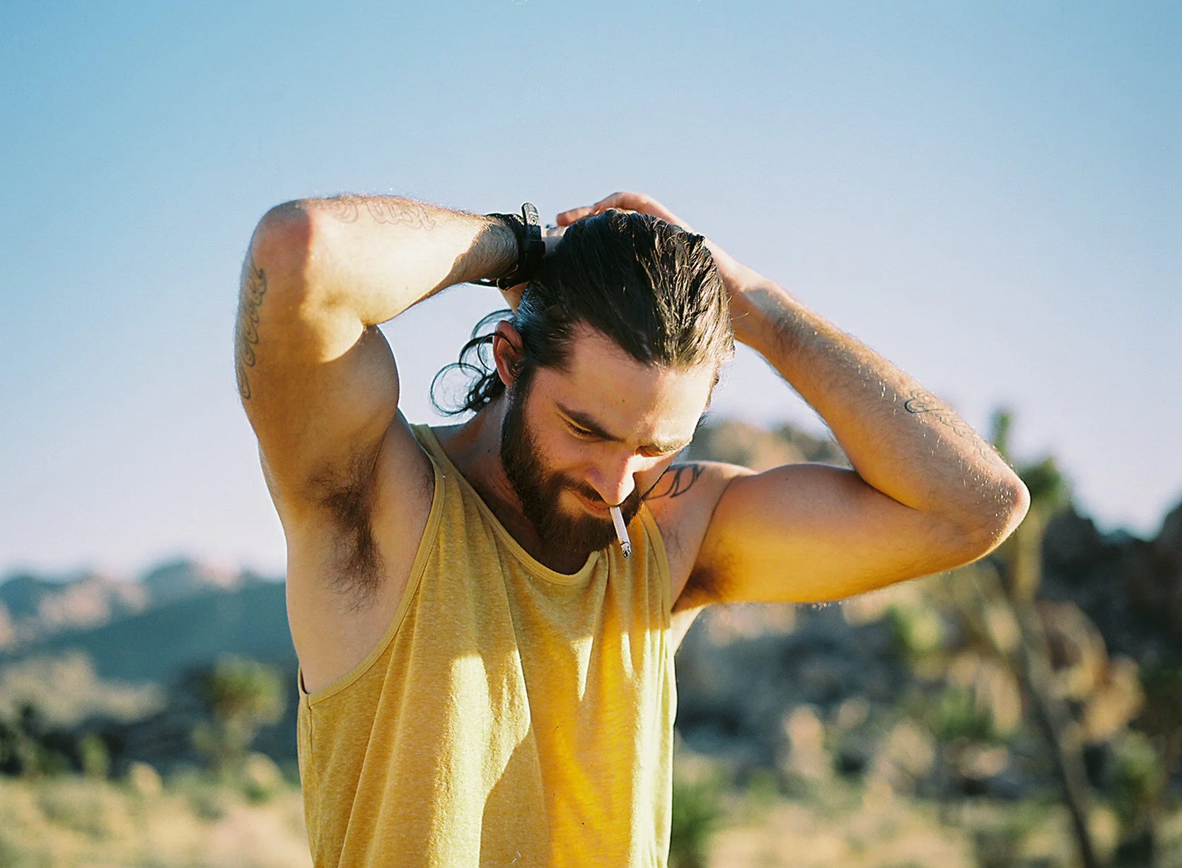 A man with long hair and a beard, wearing a yellow sleeveless shirt, standing outdoors with a sunset or sunrise sky in the background, holding his head with both hands, and a cigarette hanging from his mouth.