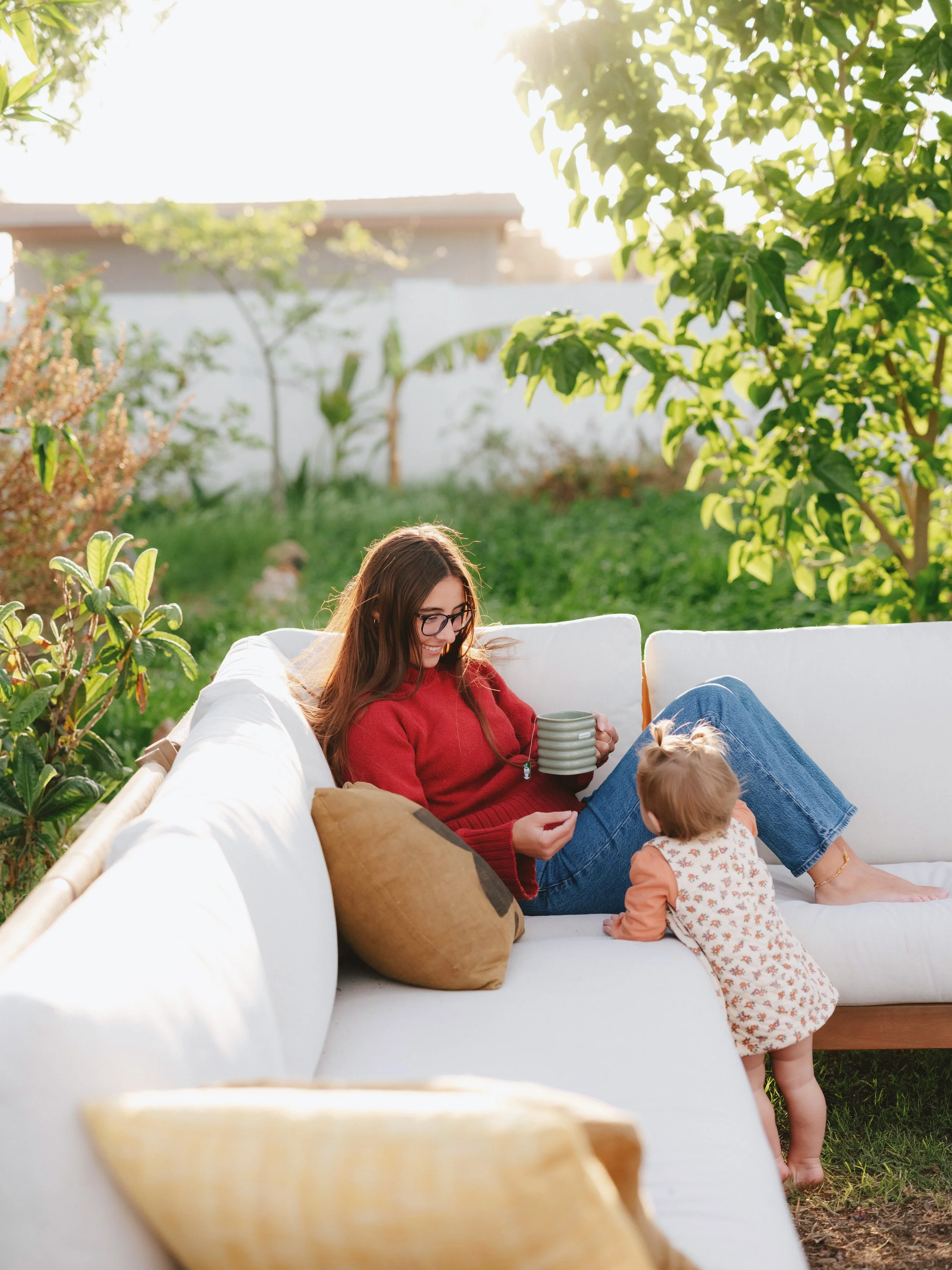 A woman and a young child sit on a white outdoor sofa in a green garden, enjoying a sunny day. The woman holds a stack of mugs, and the child stands on the sofa, reaching for the mugs.