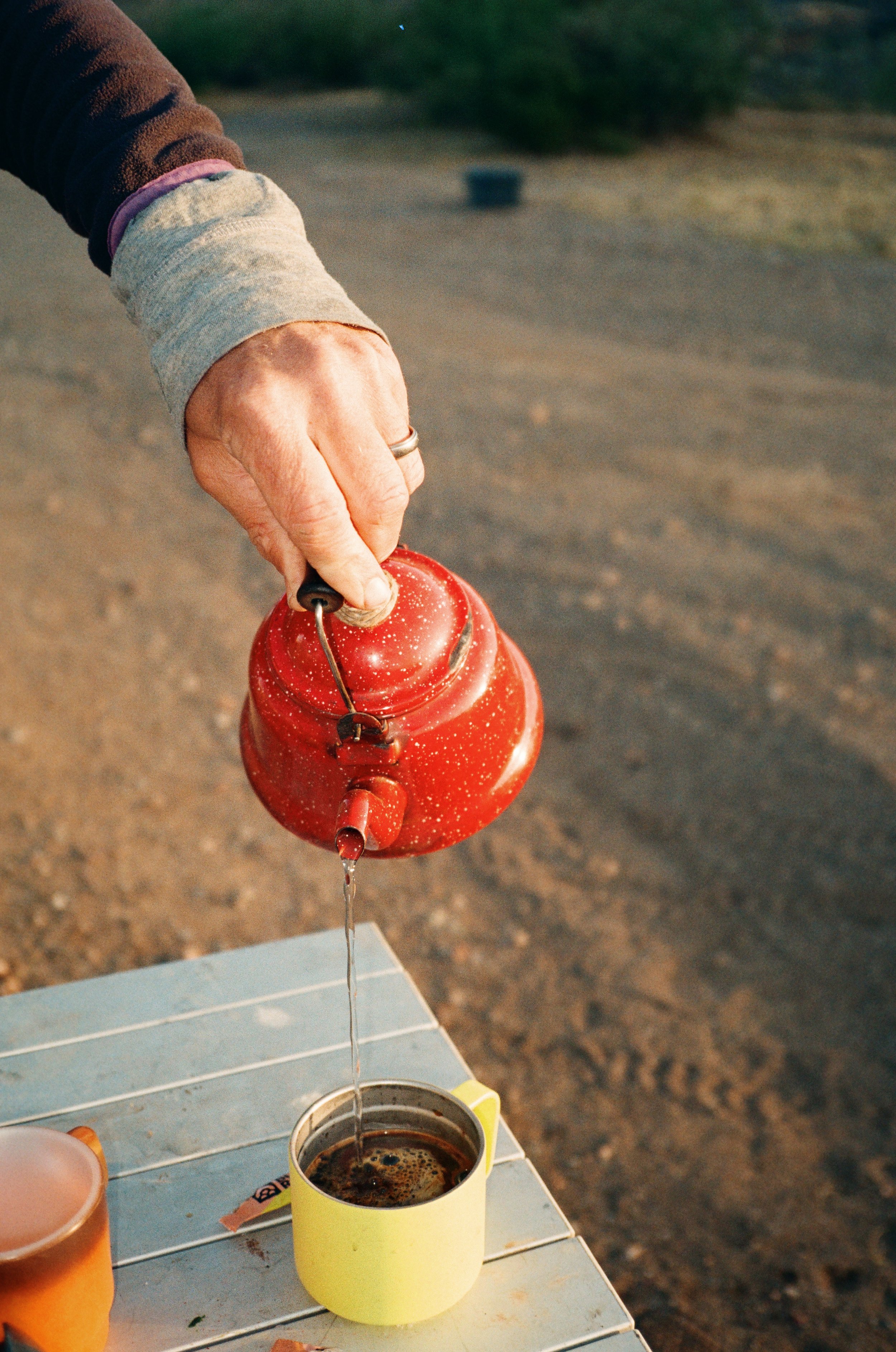 Person pouring hot water into a yellow mug containing coffee on an outdoor table.