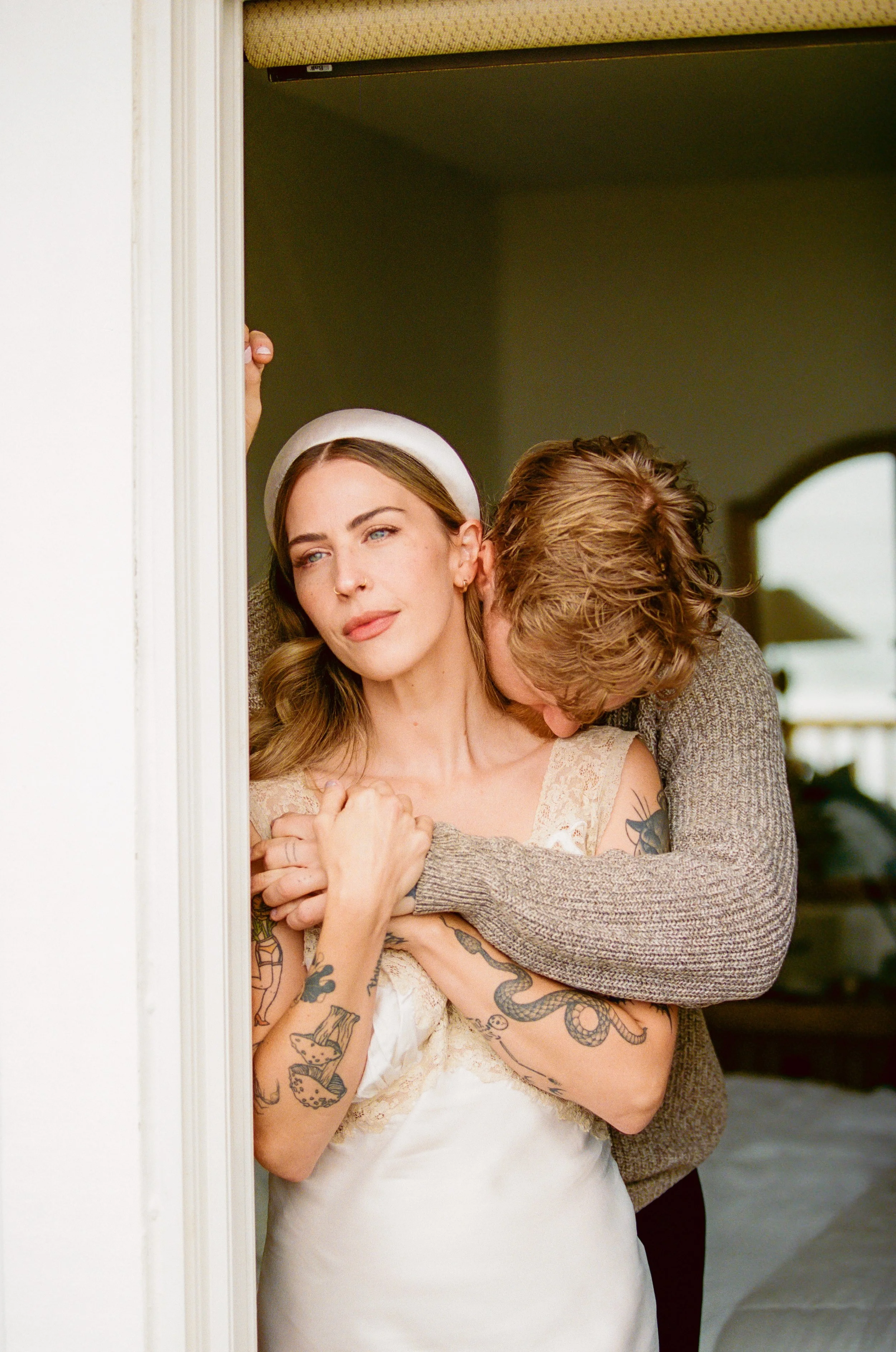 A woman with tattoos on her arms wearing a white dress and a white headband stands in a doorway, while a man embraces her from behind, whispering in her ear.
