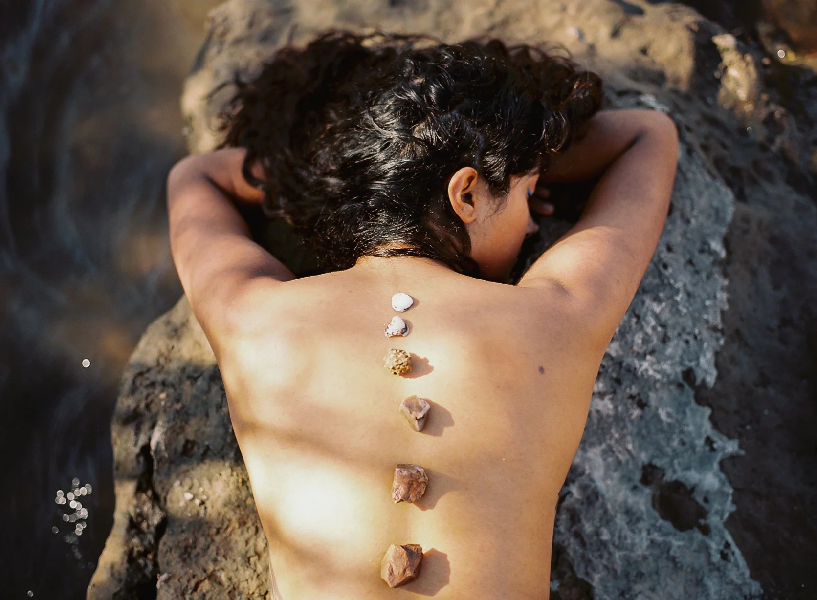A person with curly dark hair lying face down on a rock, with seven stones of different sizes and colors placed in a straight line along their back.