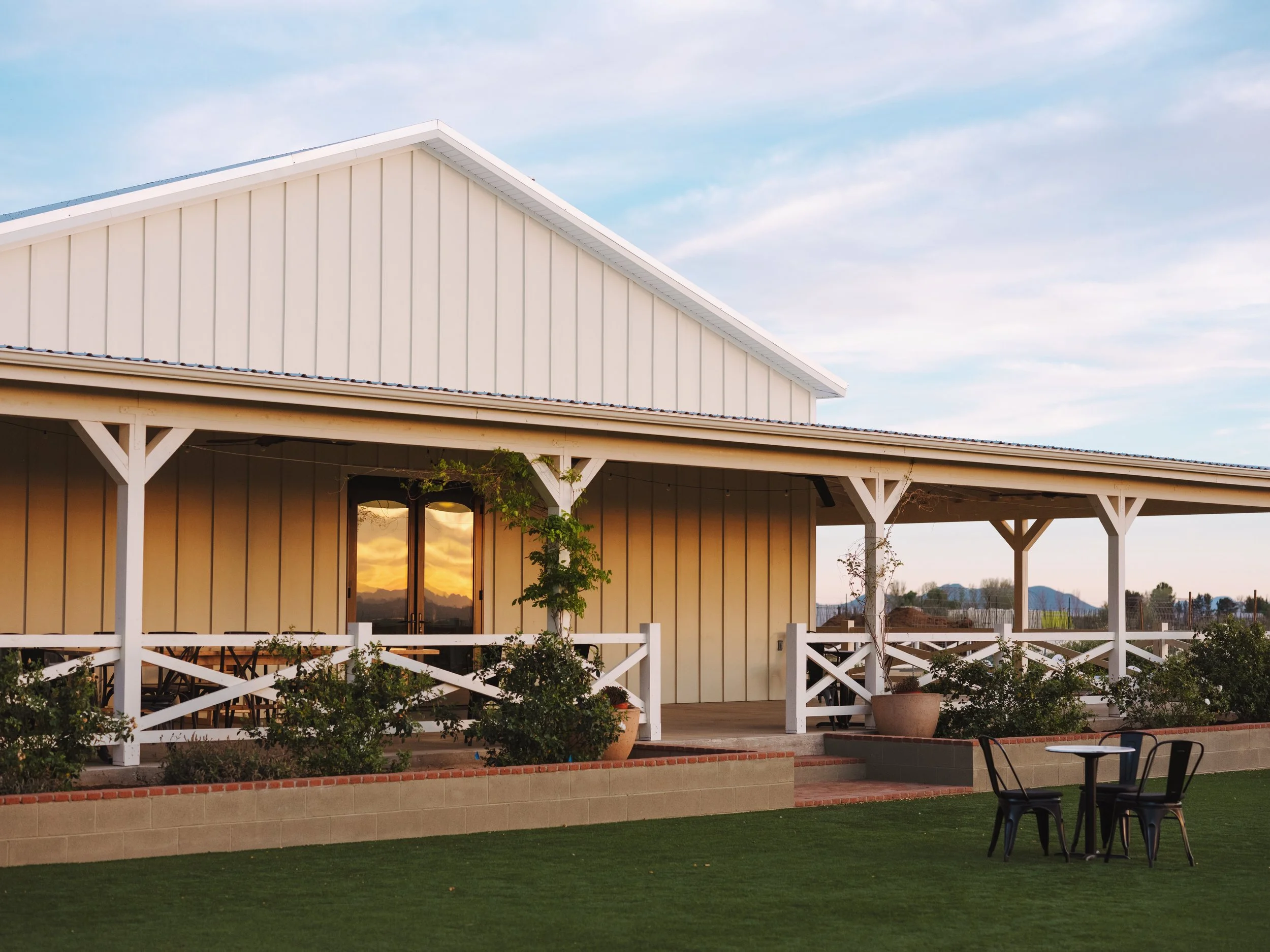 A white house with a large covered porch, surrounded by a green lawn and potted plants, with outdoor furniture and a scenic mountain view in the background during sunset.