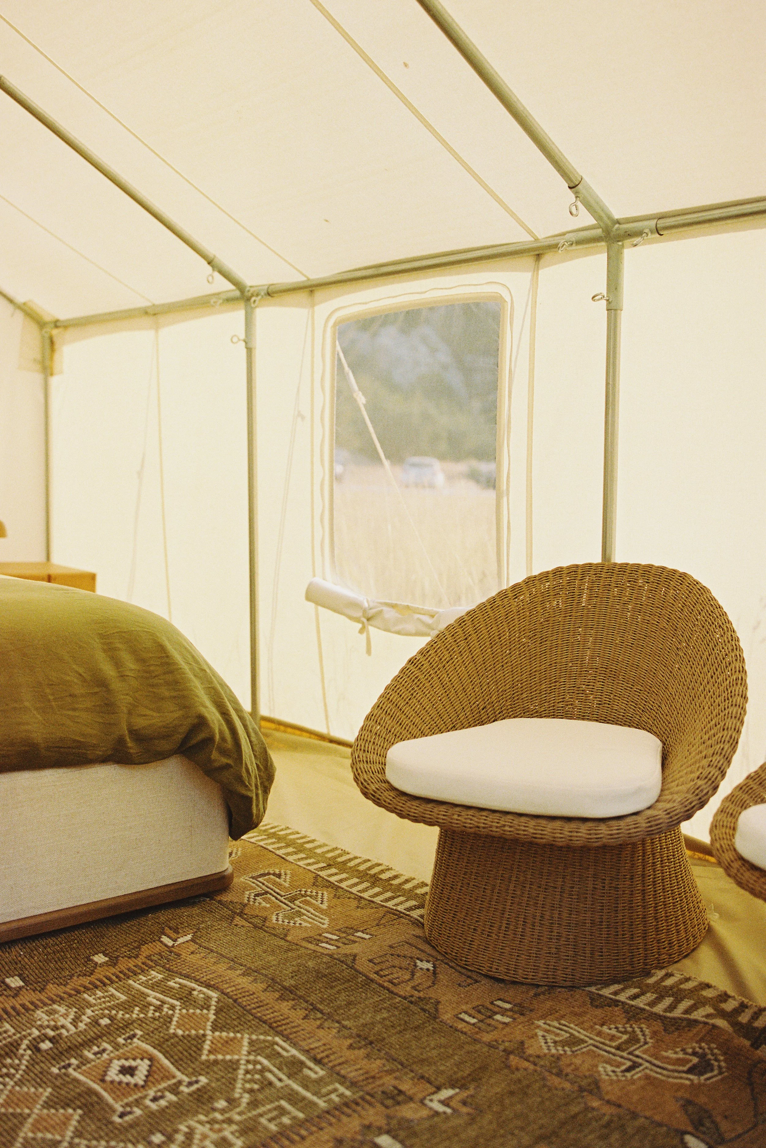 Interior of a canvas glamping tent with a wicker chair, a bed with green bedding, and a patterned rug.