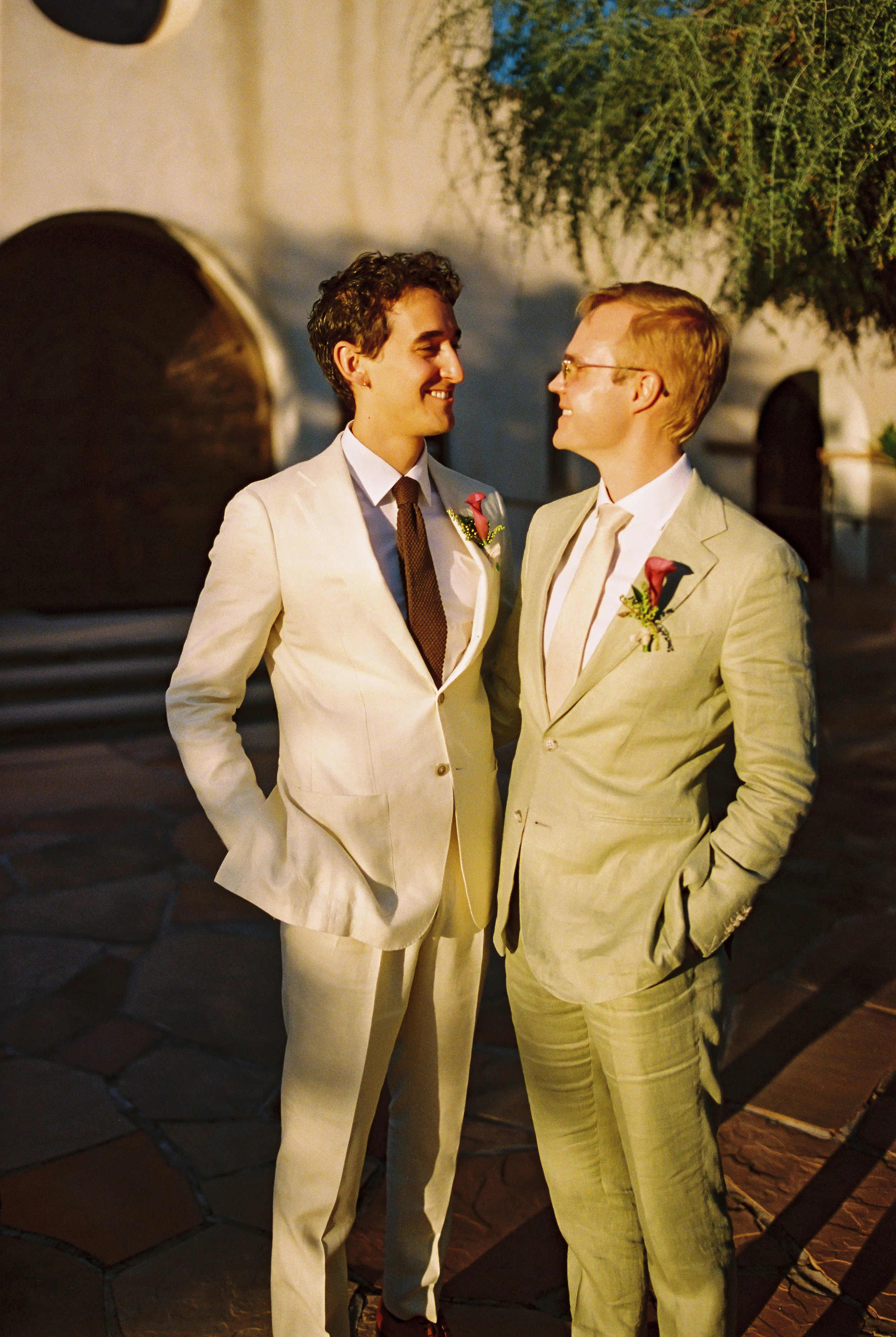 Two men in beige suits at a wedding, smiling and looking at each other, outdoors at night.