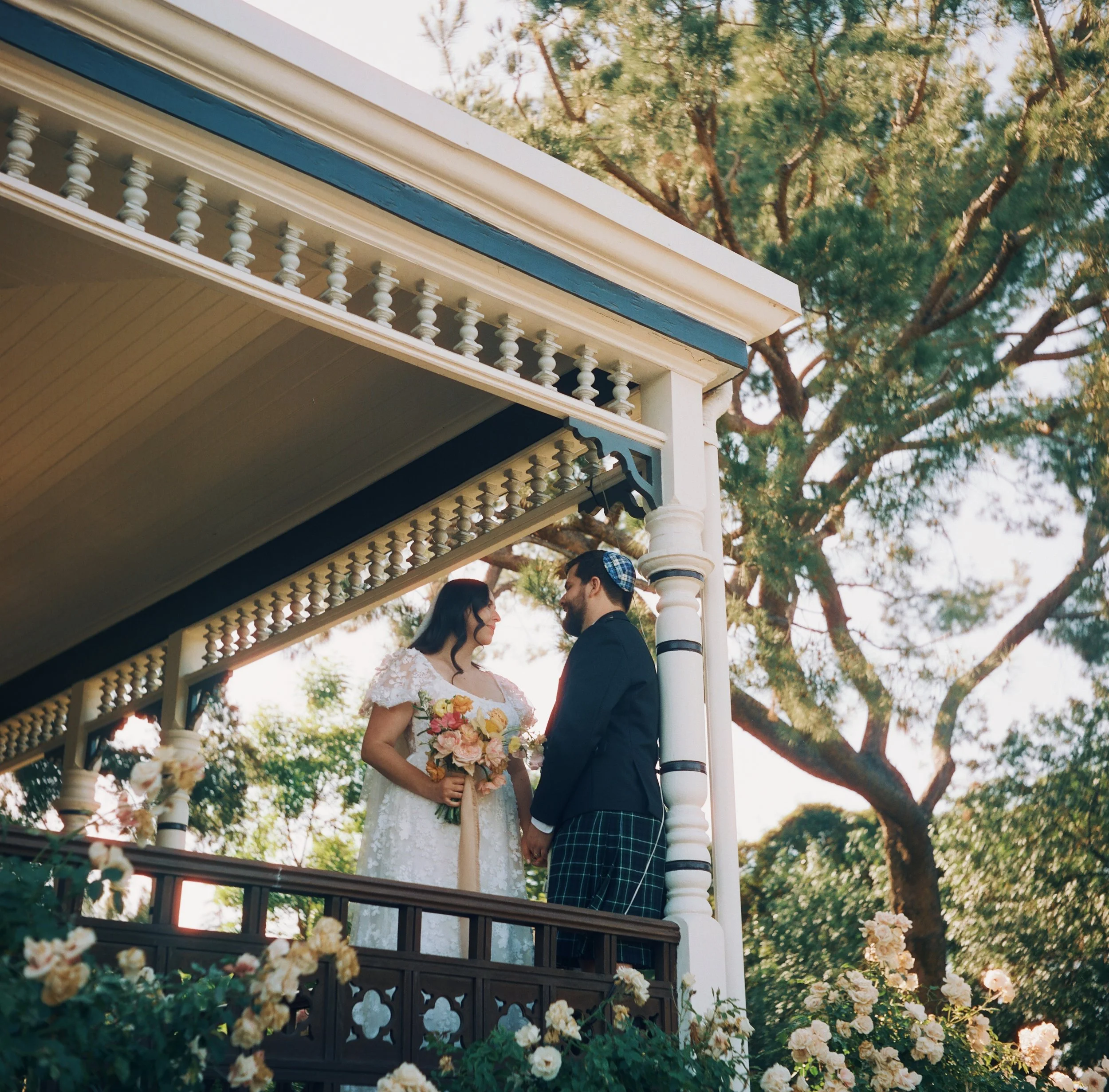 A bride and groom standing on a porch, holding hands and gazing at each other. The bride is holding a bouquet of flowers and wearing a white lace dress, while the groom is dressed in a dark suit with plaid pants and a checkered hat. The setting inclu