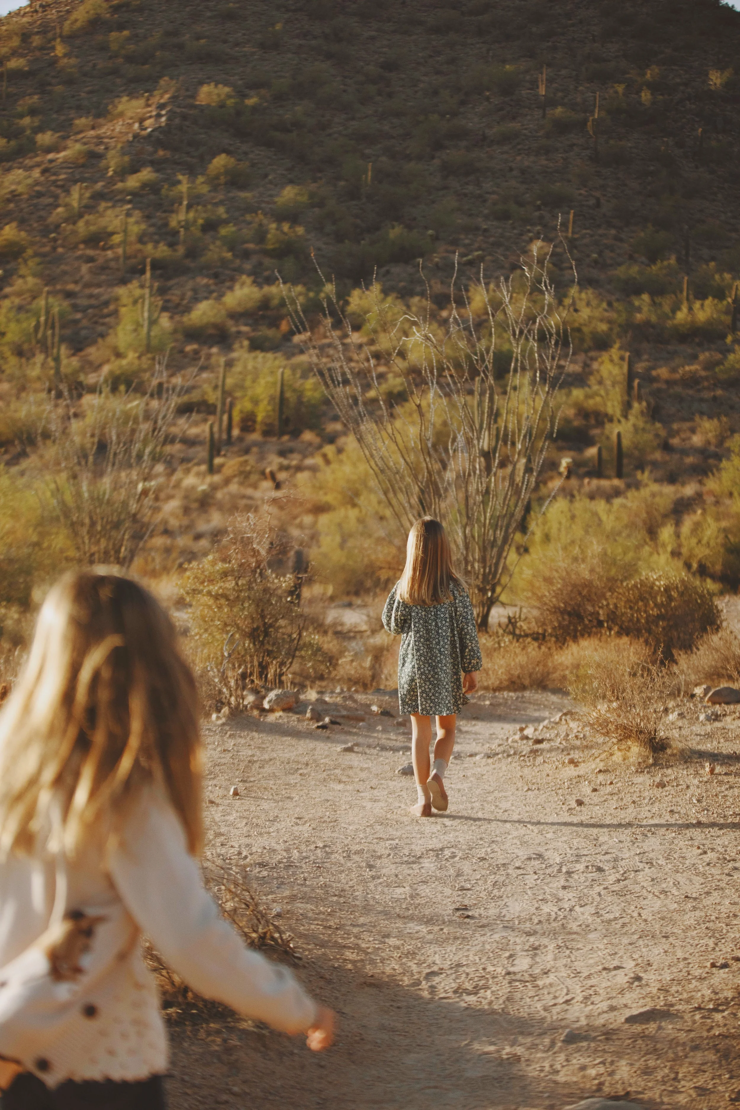 Two young girls walking on a dirt trail in a desert landscape with sparse vegetation, cacti, and hills in the background during sunset.