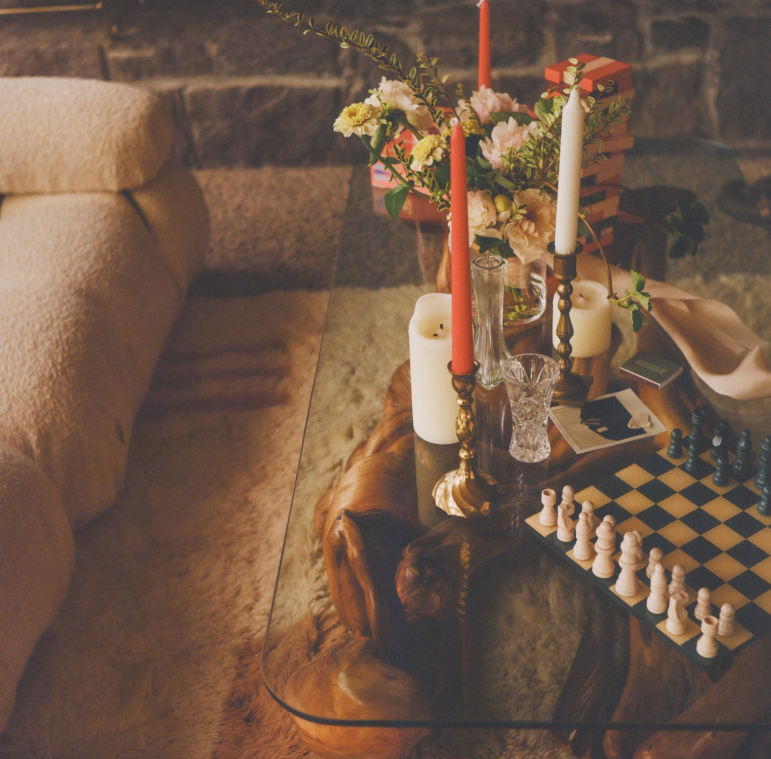 A cozy living room with a glass-topped wooden coffee table, decorative candles, a vase of flowers, a chessboard, and a plush beige sofa against a brick wall.