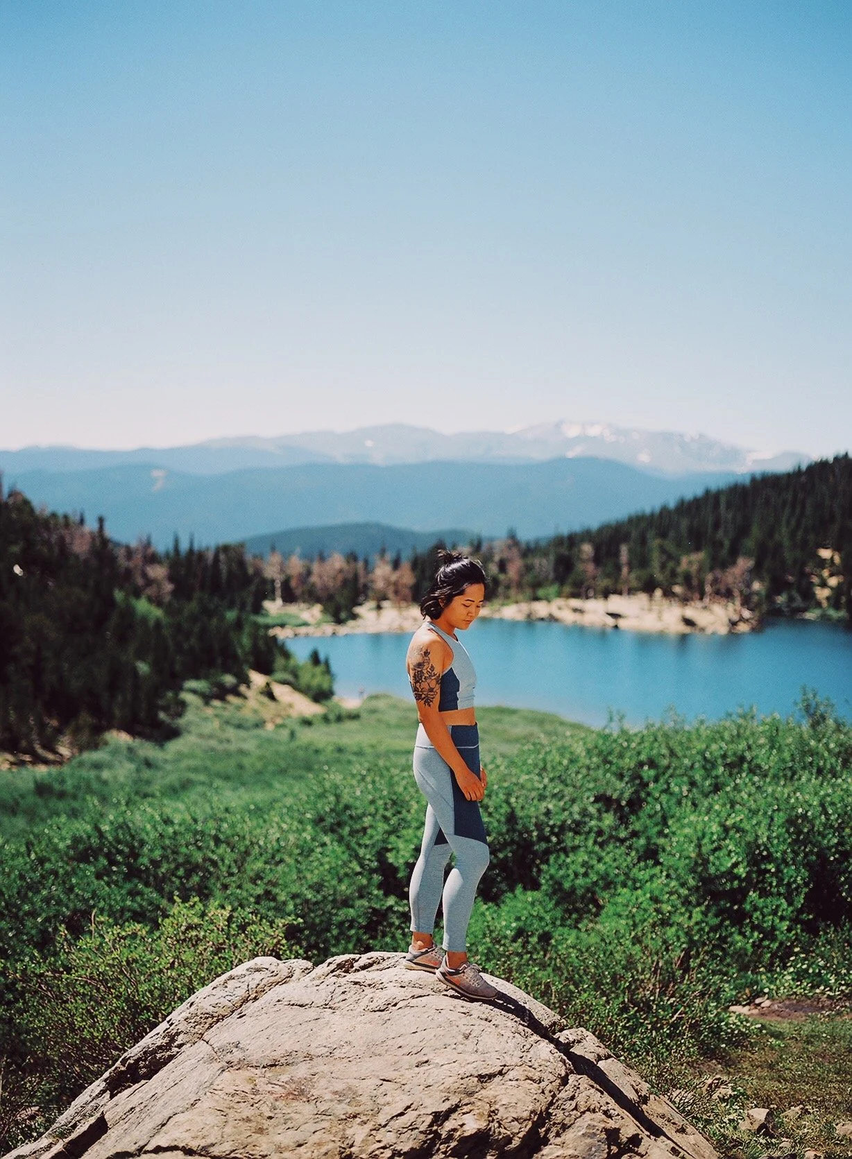 A woman in athletic clothing standing on a large rock outdoors, overlooking a lake with mountains in the distance on a clear, sunny day.