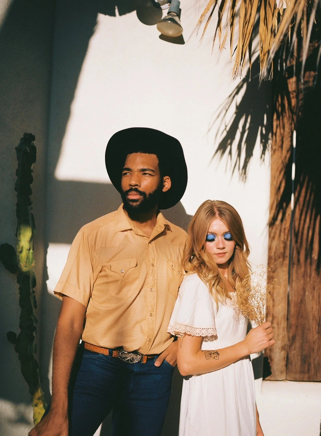 A man with a beard and black hat, and a woman with blonde hair and blue eye makeup, standing in front of a light-colored wall decorated with a cactus and shadow of palm leaves. The woman holds dried flowers.