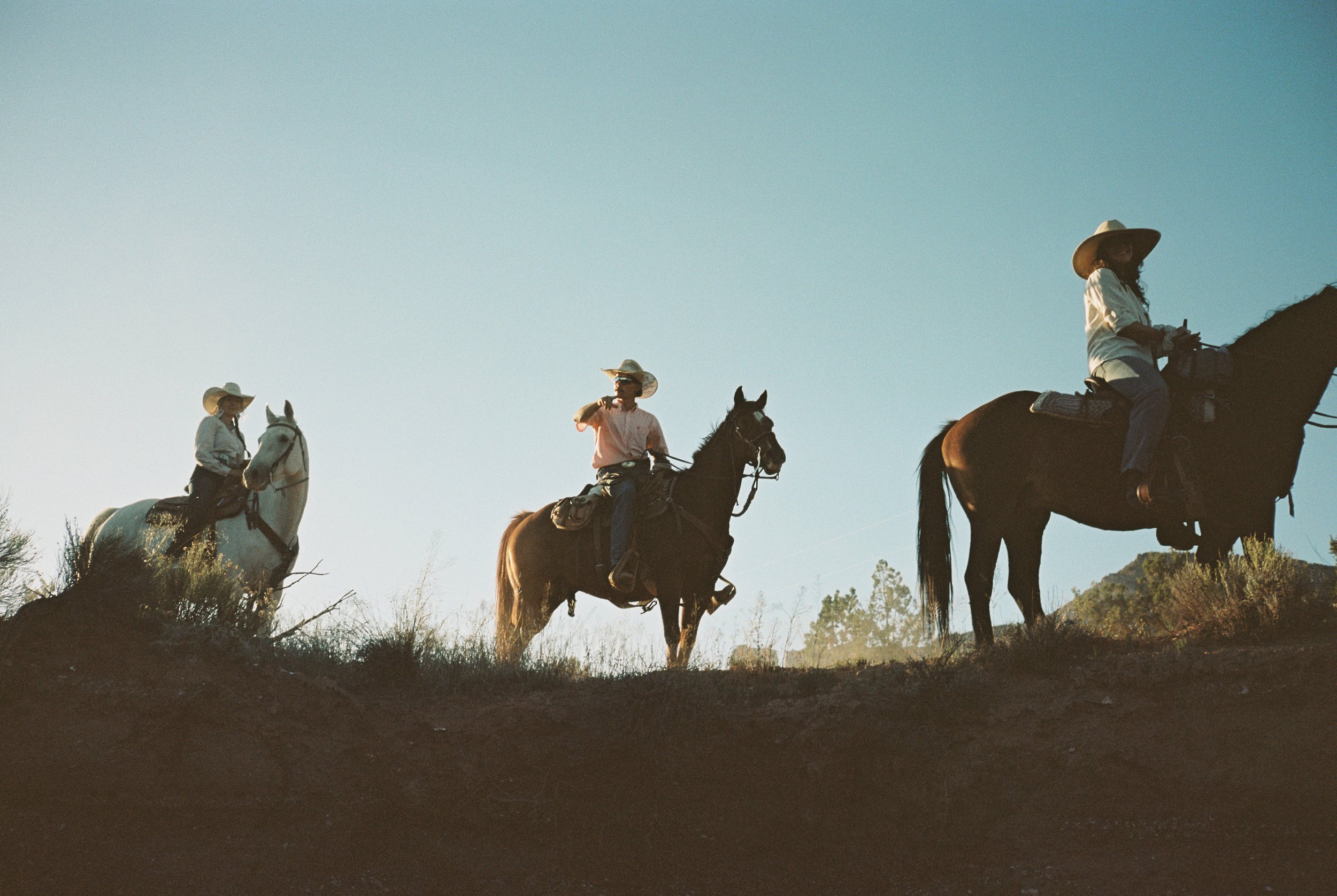 Three horseback riders with cowboy hats on a dirt trail during sunset.