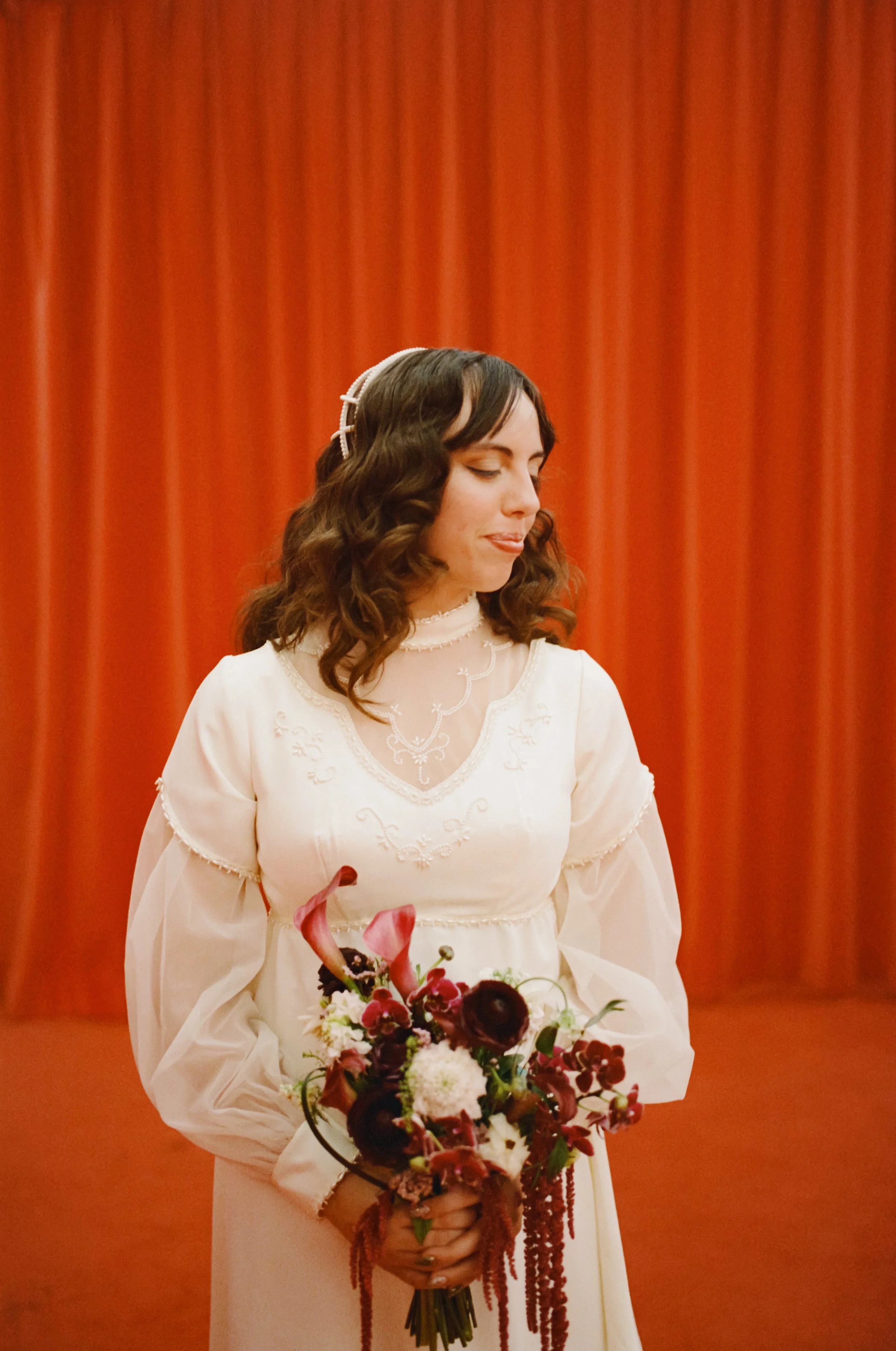 A woman in a vintage white dress with lace details, holding a colorful bouquet of flowers, with a red curtain in the background.