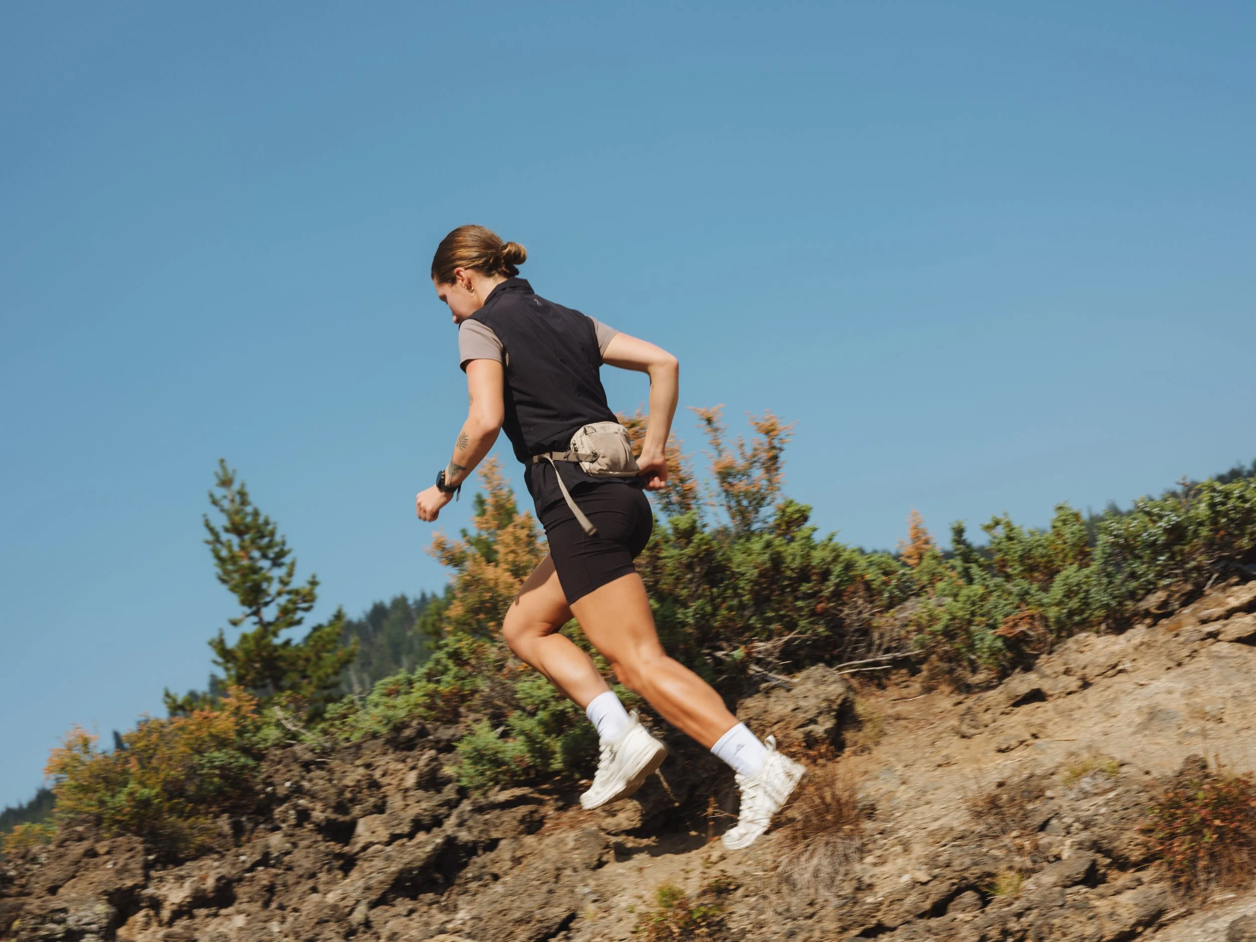 A woman running uphill on a trail with rocky terrain, greenery, and trees in the background under a clear blue sky.