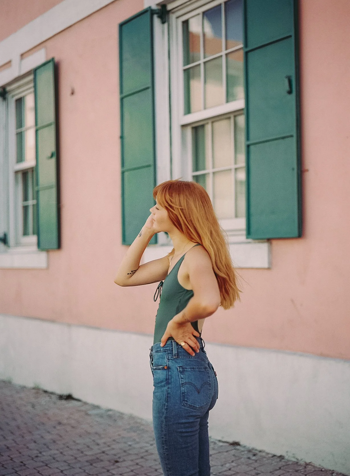 A woman with red hair, wearing a green tank top and blue jeans, standing outside with a pink building and green window shutters in the background.