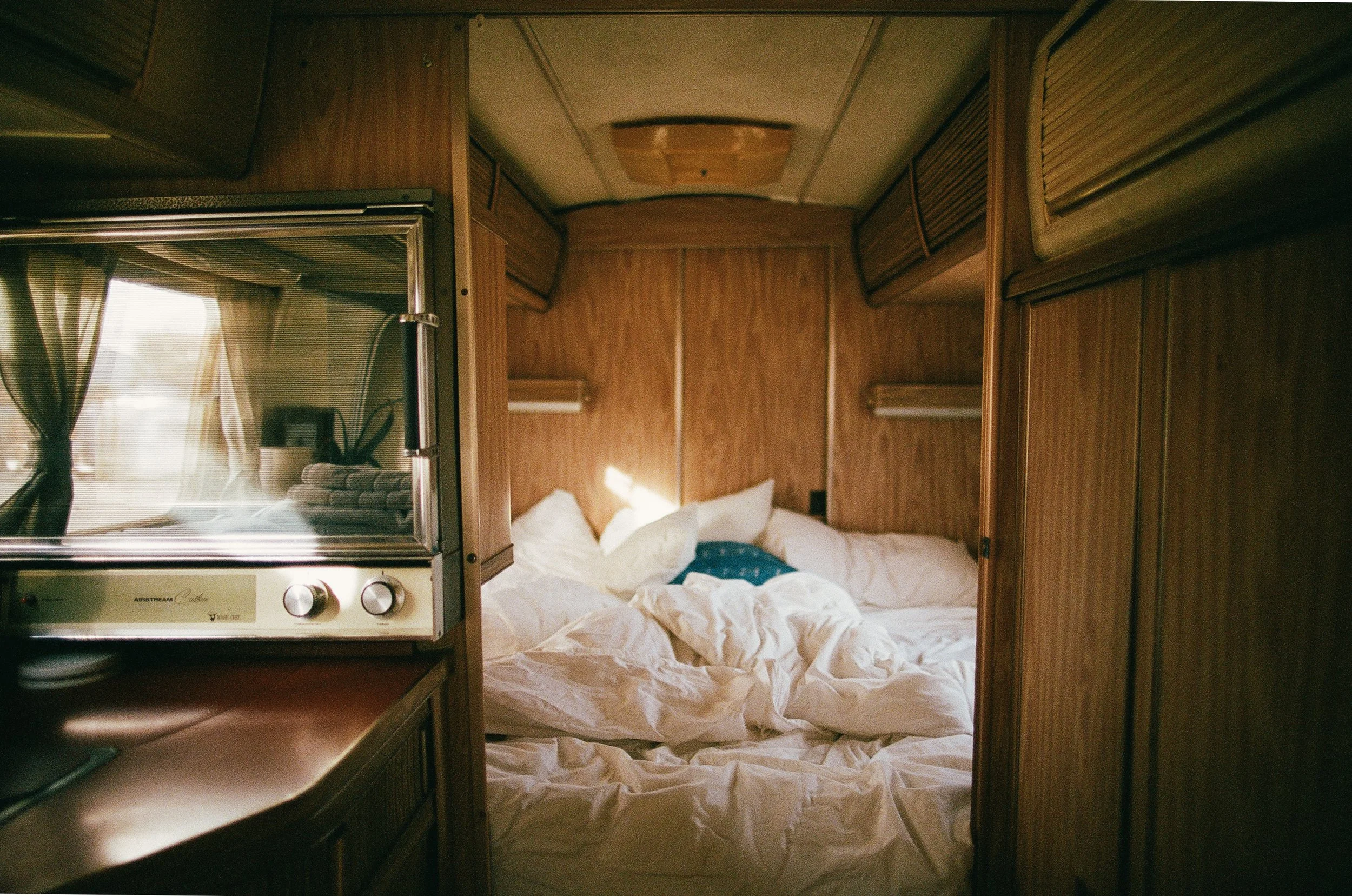 A cozy, messy bed inside a small wooden camper or cabin, with white rumpled sheets and pillows, and sunlight streaming through a window.
