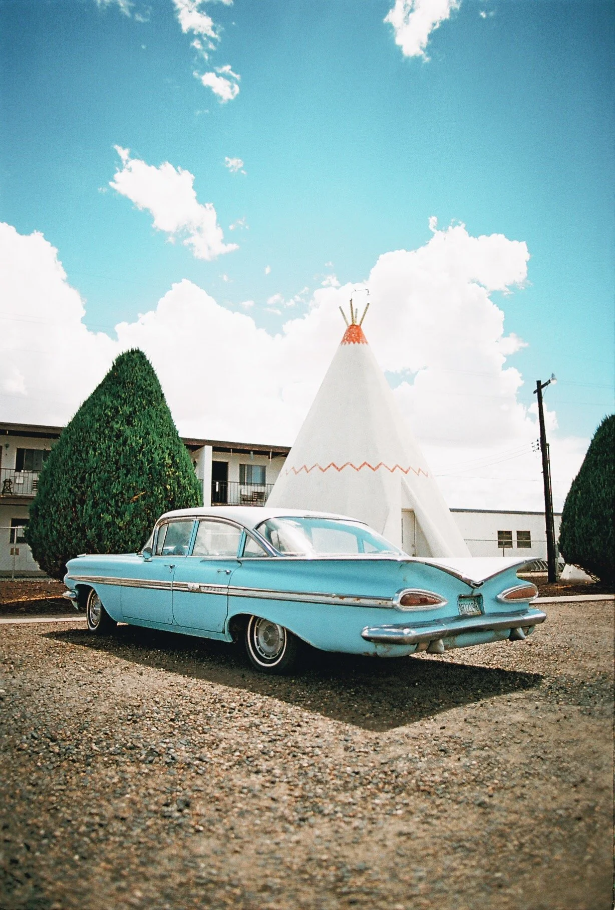 A vintage blue car parked in front of a white teepee tent with a red zigzag pattern, flanked by neatly trimmed bushes, under a partly cloudy sky.