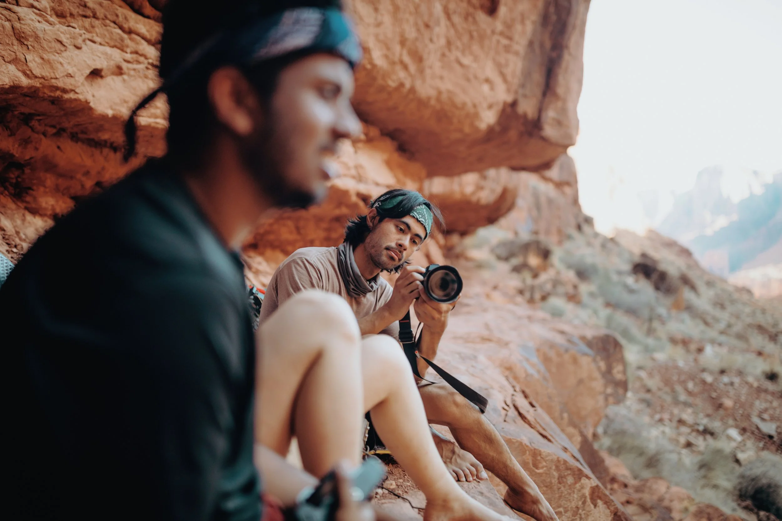 Two young men sitting on a rocky ledge in a desert landscape, taking a break. One man in the background, holding a camera and looking at the other man, who is in the foreground and out of focus.