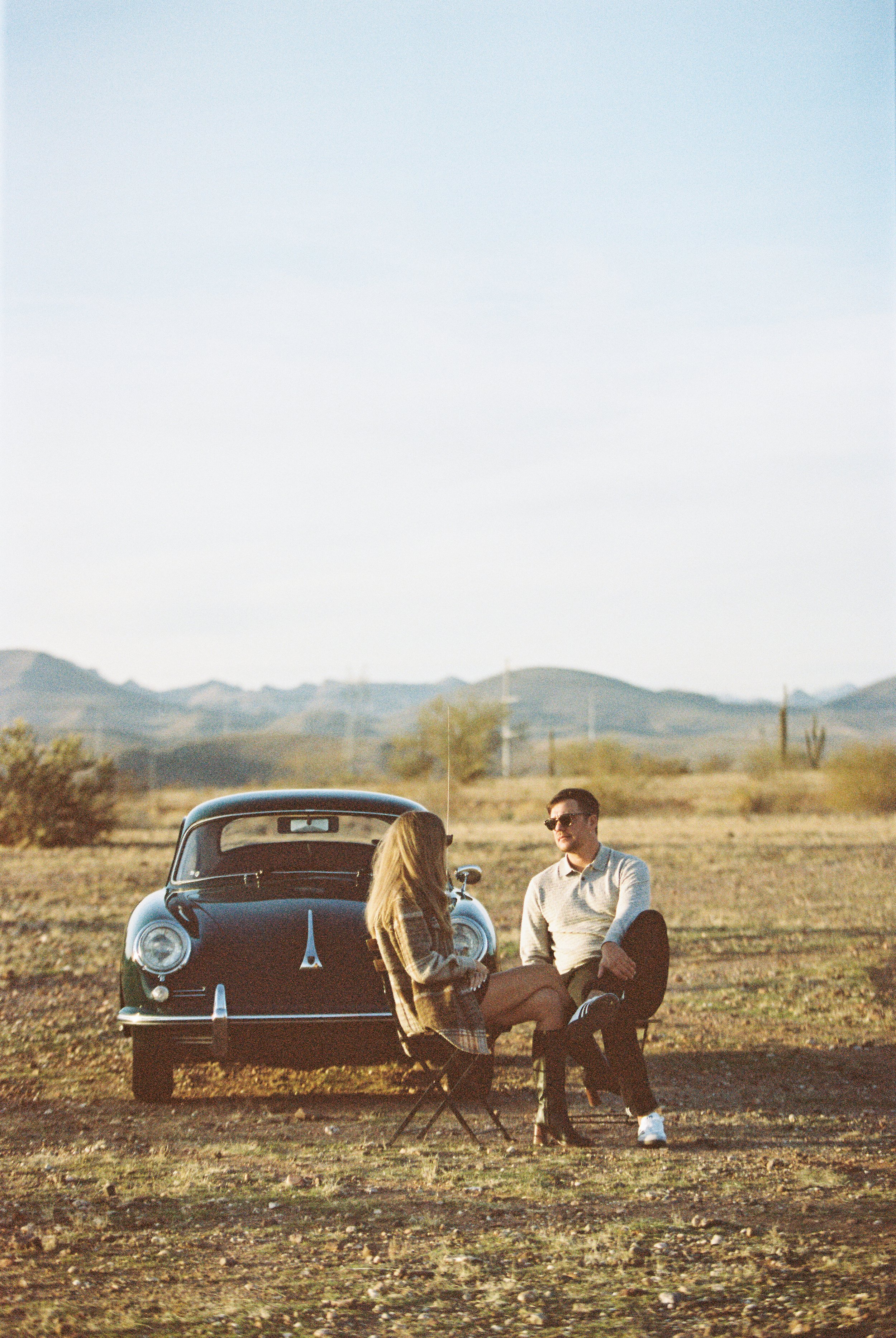 A man and woman sitting and talking outdoors next to a vintage black car in a desert landscape with mountains in the background during sunset.
