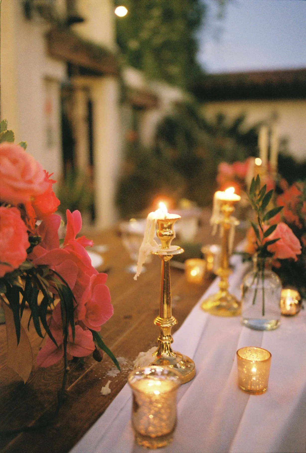 A beautifully decorated outdoor dinner table with pink flowers, gold candle holders with lit candles, and small votive candles, set during the evening with soft lighting.