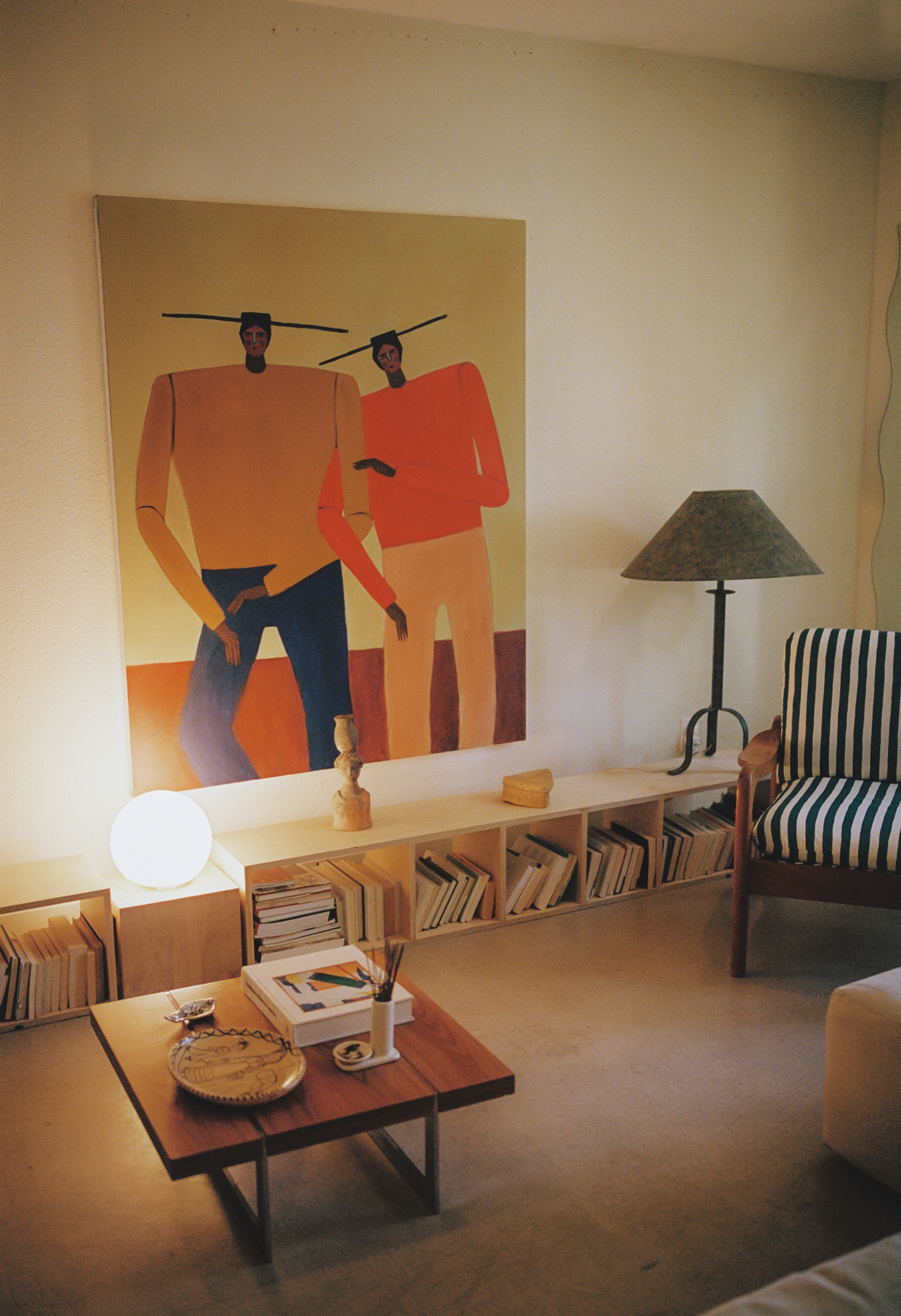 Living room with a large, colorful painting of two elongated figures, a white spherical lamp, a wooden coffee table with art supplies, a striped armchair, a standing lamp, and a bookshelf with books.