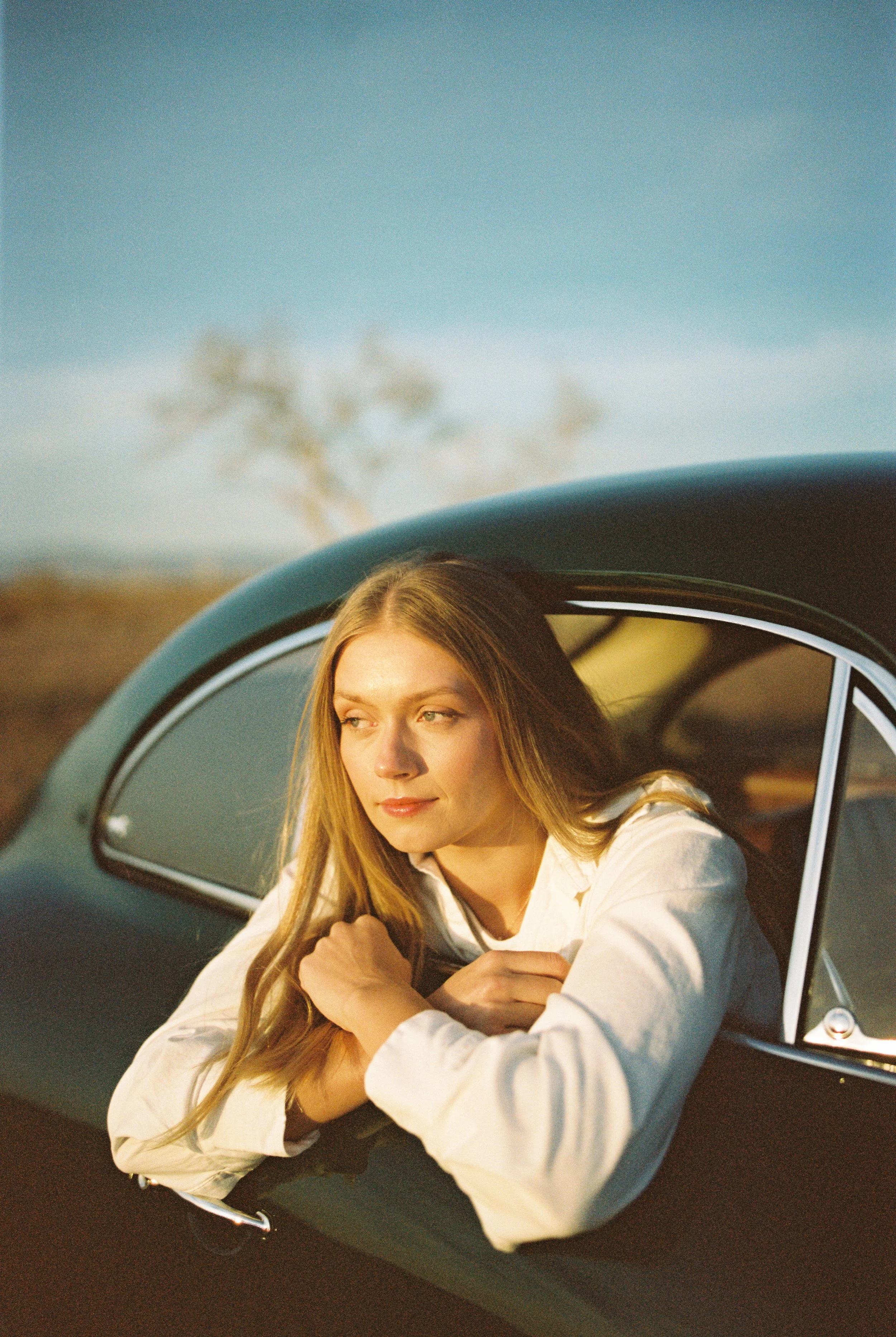 A woman with long blonde hair leaning out of a vintage black car window, looking into the distance during sunset with a soft natural light, in an open outdoor setting.