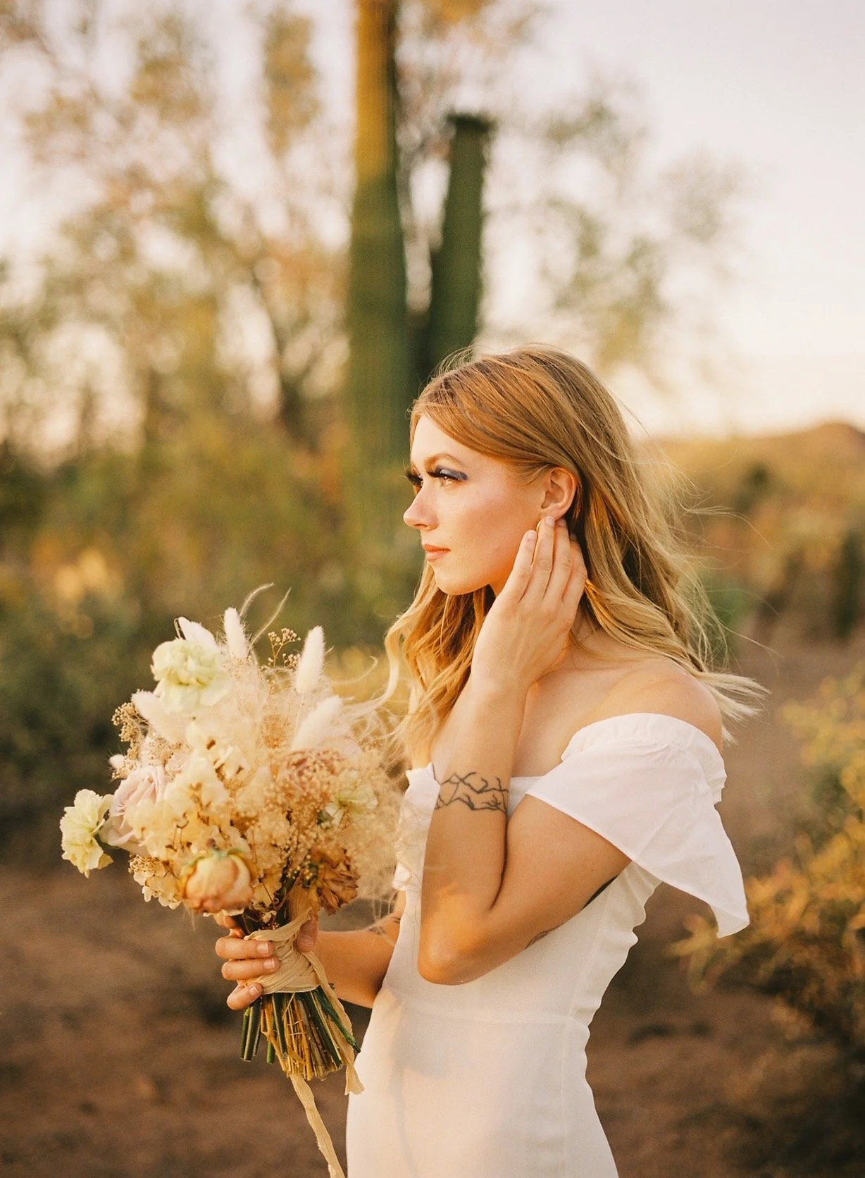 A woman with red hair in a white dress holding a bouquet of flowers outdoors in a desert landscape with cacti, during sunset.