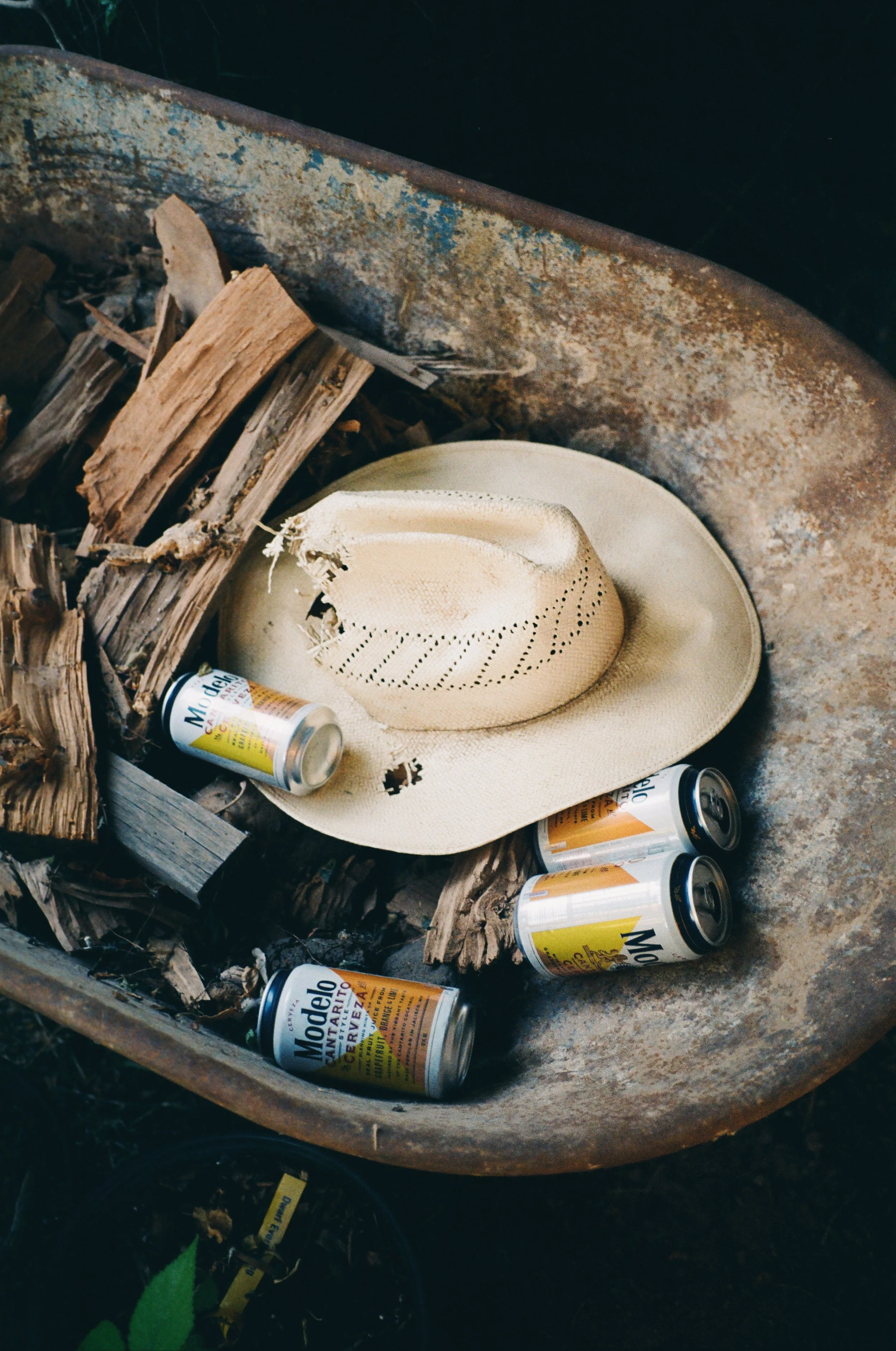 A worn cowboy hat, four empty Modelo beer cans, and a fire pit with partially burned wood inside.