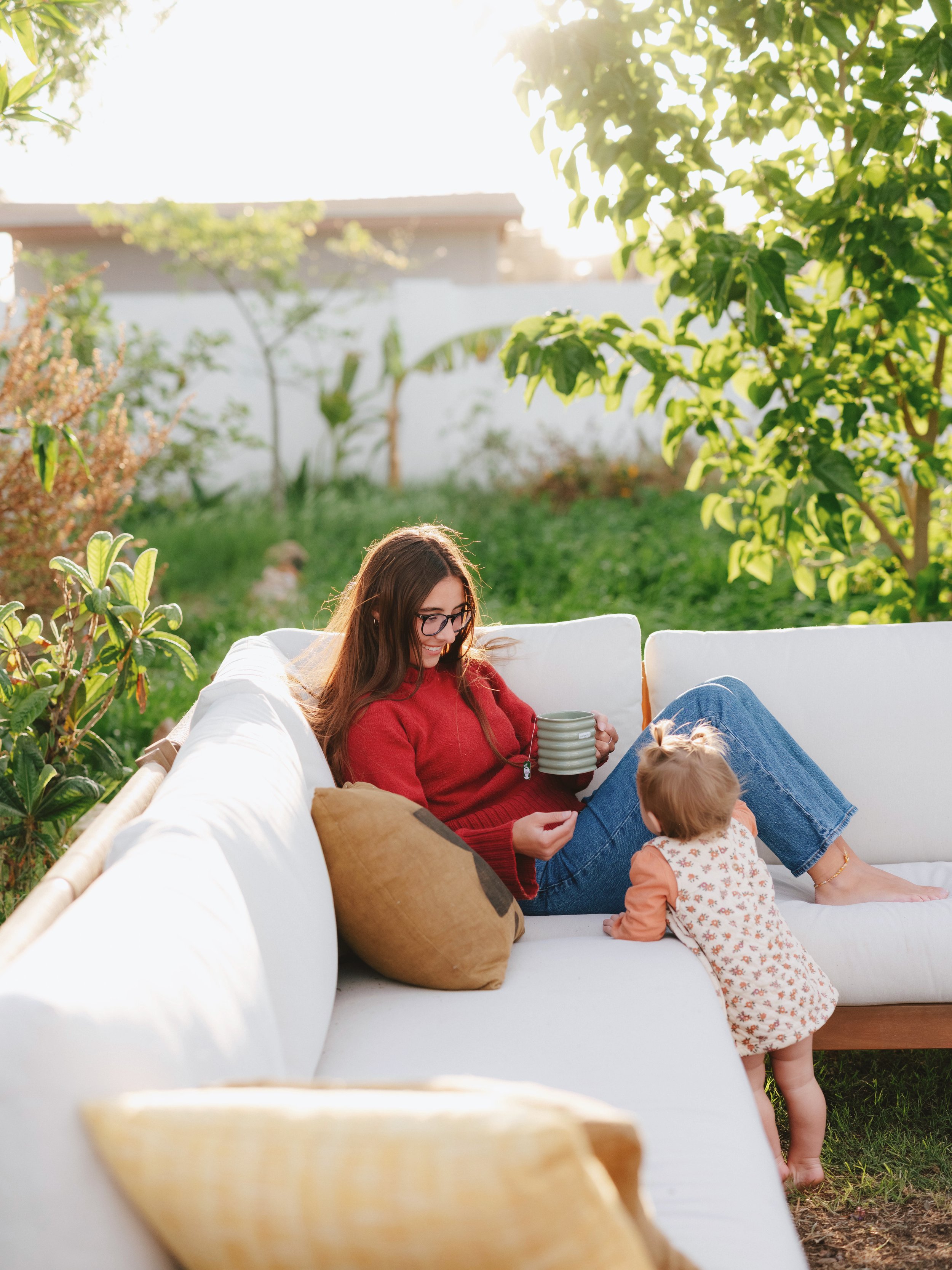 A woman with glasses and a red sweater sitting on a white outdoor sofa, holding colorful cups, while a small child in a patterned dress stands nearby in a lush garden during daytime.