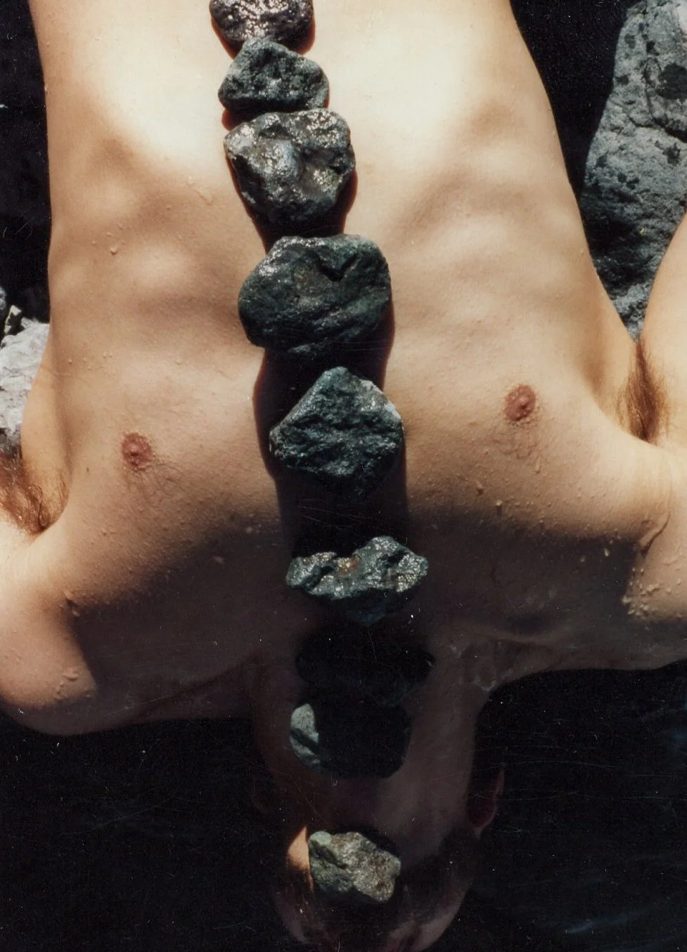 A person is submerged in water with their face, chin, and chest visible. They are holding a vertical line of six dark rocks, which are reflected in the water below them.