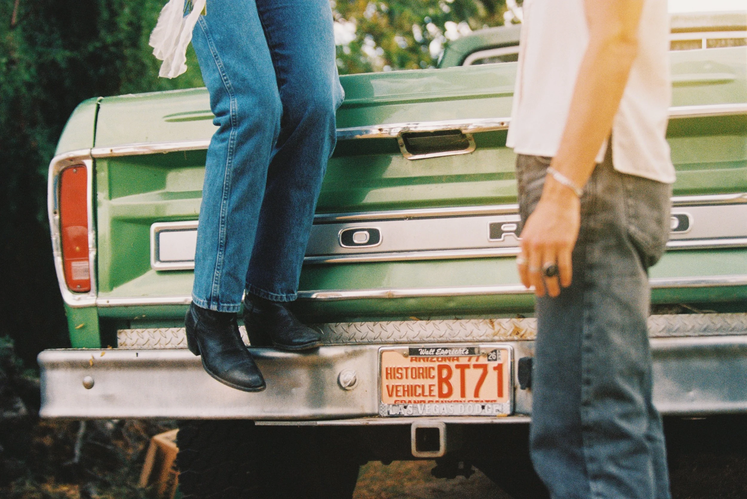 A person wearing black cowboy boots and blue jeans sitting on the tailgate of a vintage green Ford pickup truck, with another person standing nearby. The truck has a distribution license plate from Nevada indicating it is a historic vehicle.