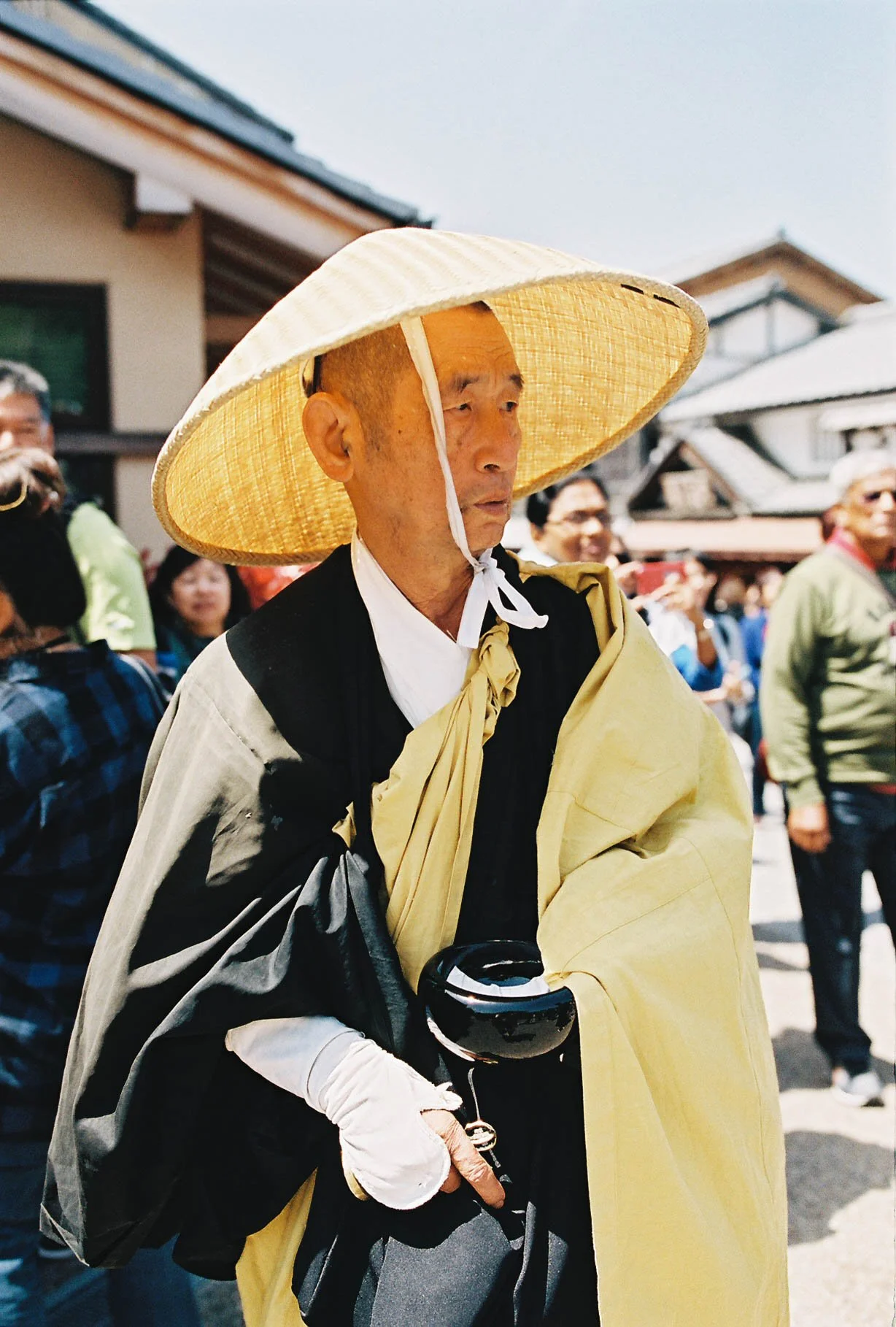 An elderly man in traditional Asian attire, wearing a large straw hat and holding a black bowl, stands among a crowd of people outdoors with traditional-style buildings in the background.