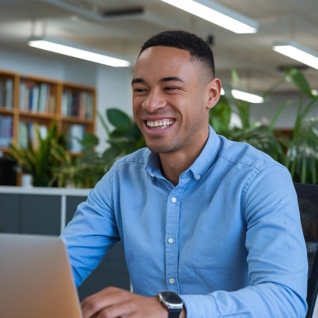 A cheerful male office worker smiling while working on his laptop in a bright and modern office space. This image highlights "Get DeskFit" programs designed to enhance workplace productivity and employee satisfaction.