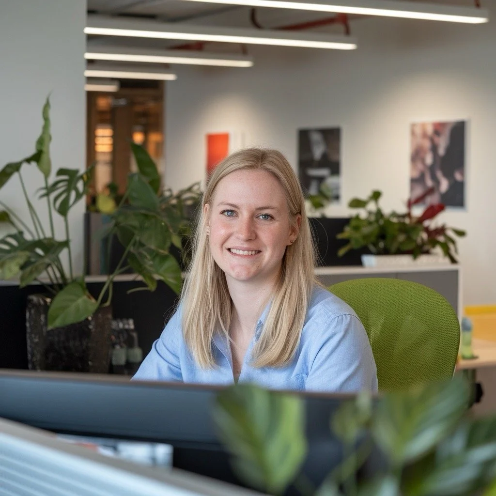 A blonde office worker smiling while sitting at her desk, surrounded by greenery in a modern workplace. This image emphasizes "Get DeskFit" solutions for boosting employee wellness and fostering a healthier work culture.
