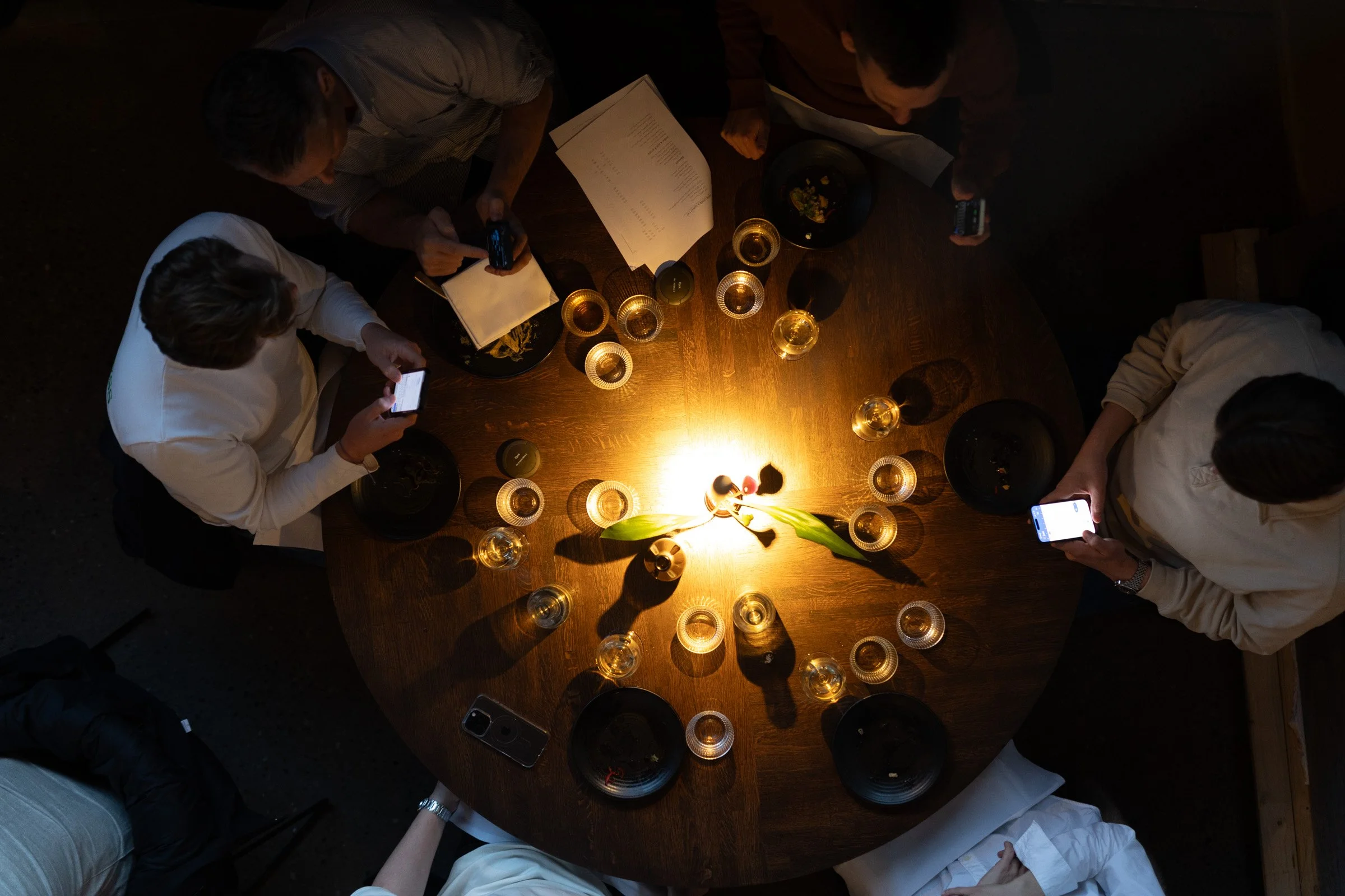 Overhead bilde av seks personer rundt et rundt bord med lys og drinker, mens de bruker mobiltelefoner.
