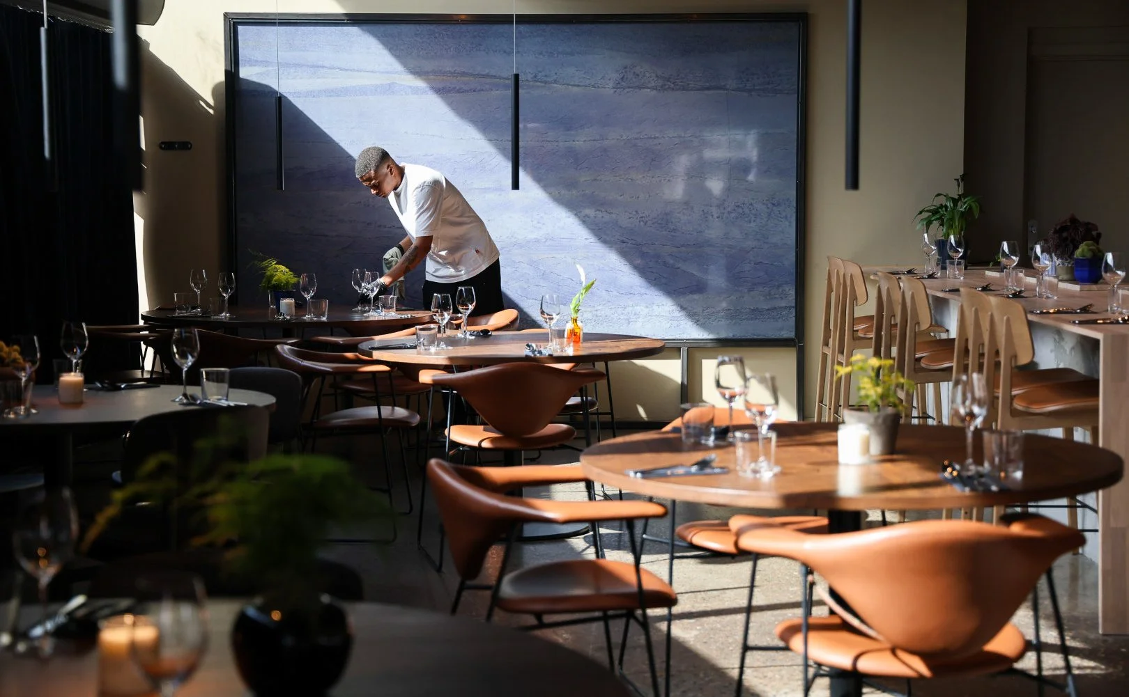 En person dekker bord i et restaurant med flere tomme bord og glass, sollys skinner inn gjennom et vindu.